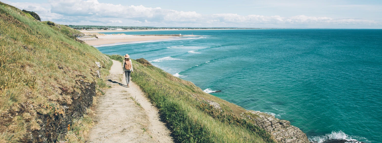 Hiker, GR223, grass, cliff path along the blue sea, blue sky, sandy beach in the distance - © Maxime Coquard for Best Jobers - Attitude Manche