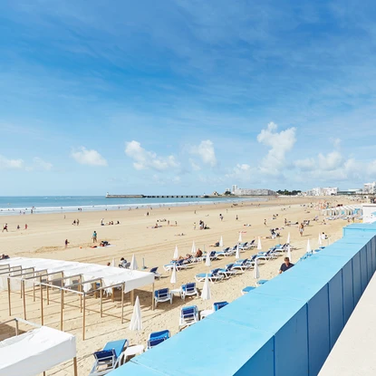 Remblai des Sables d’Olonne sous un beau ciel bleu, avec vue sur la grande plage de sable fin, la pendule en second plan et la jetée au loin.