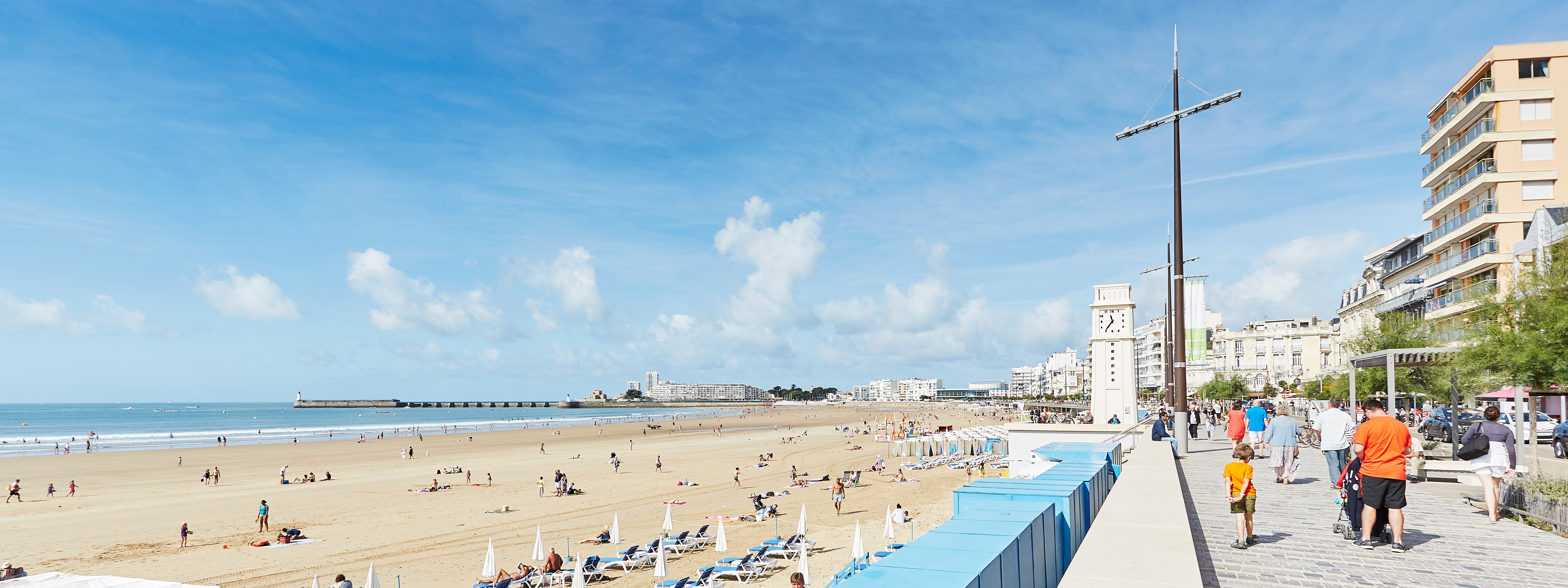 Remblai des Sables d’Olonne sous un beau ciel bleu, avec vue sur la grande plage de sable fin, la pendule en second plan et la jetée au loin.