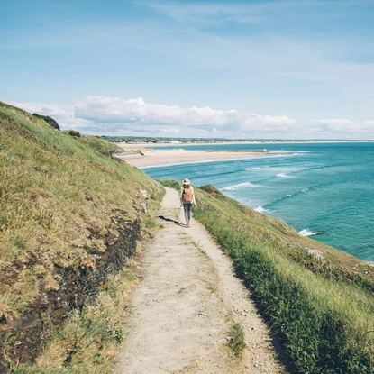 Randonneuse, GR223, herbe, chemin sur falaise longeant la mer bleue, ciel bleu, plage sable au loin