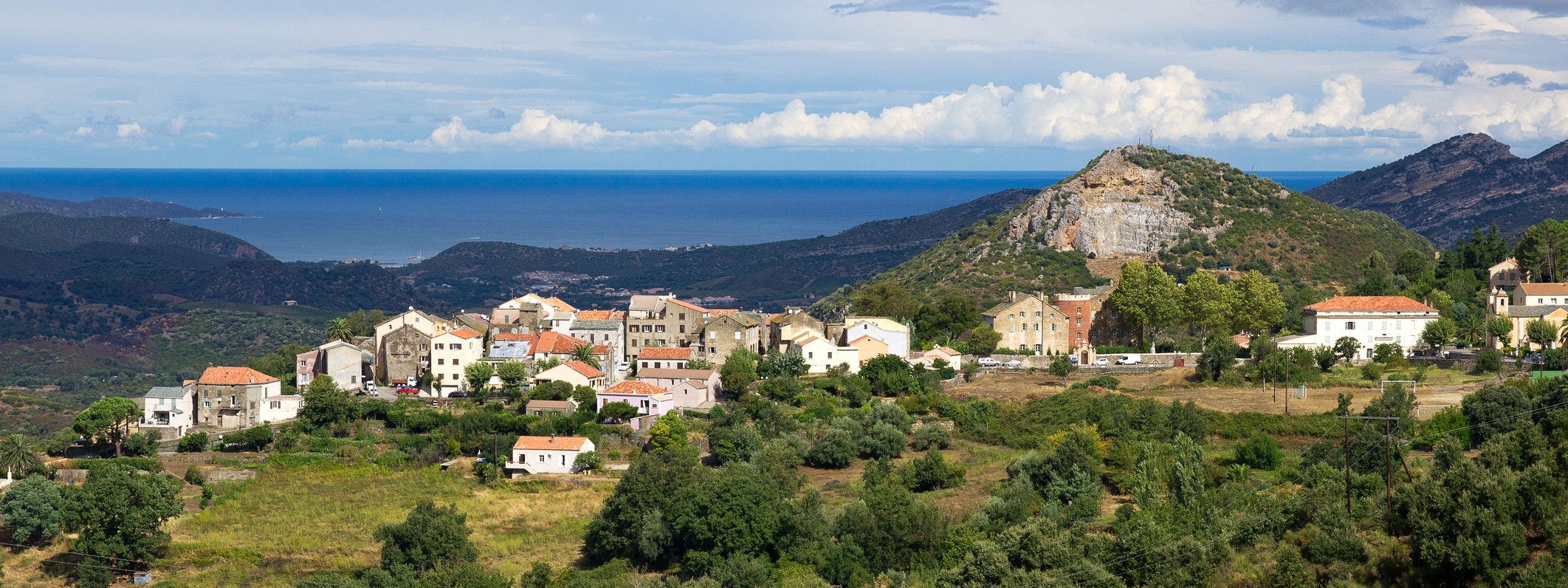 Maisons du village d'Oletta nichées dans les collines corses avec panorama sur la campagne du Nebbiu et la mer au loin.