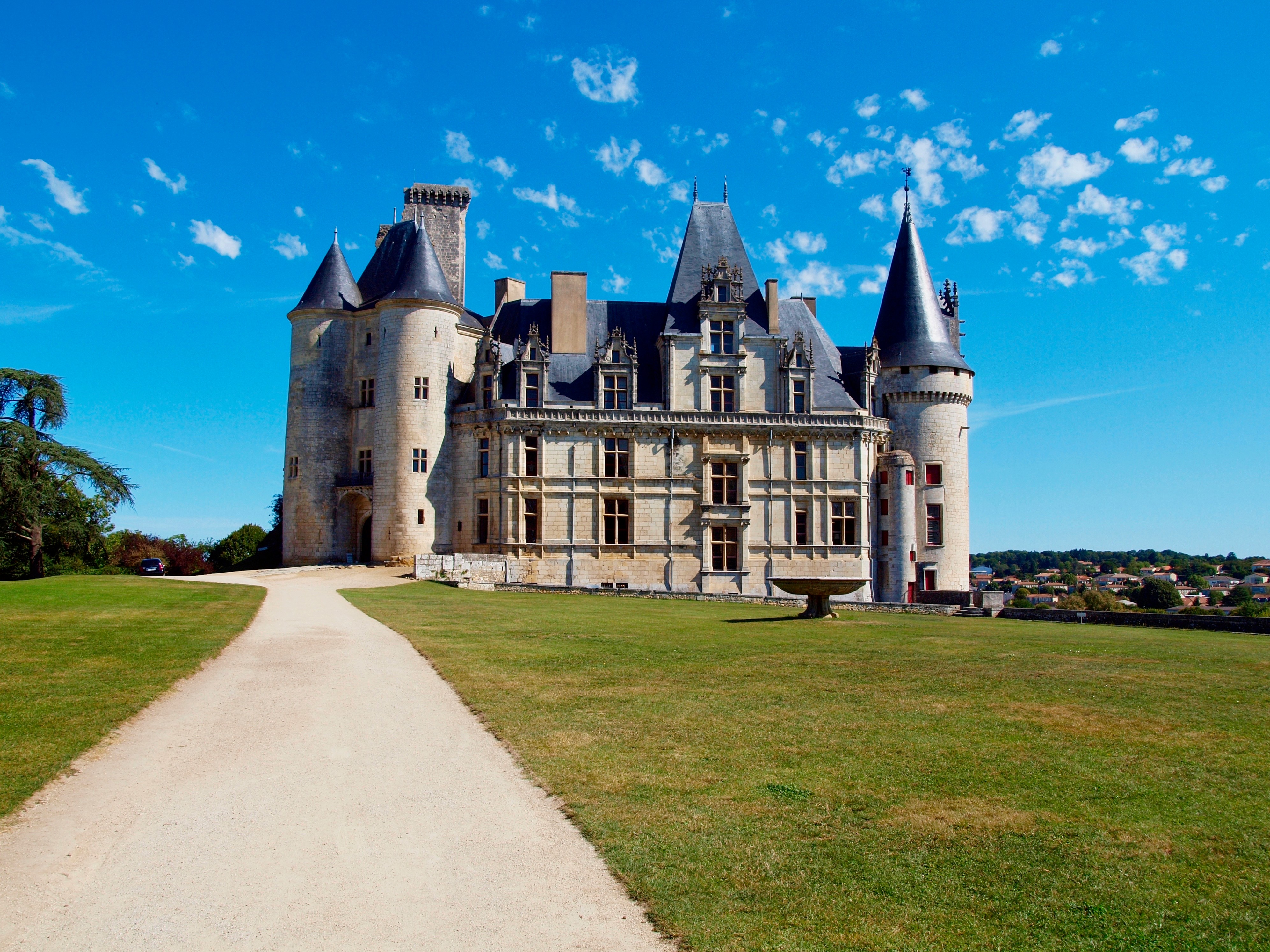 Depuis un chemin à côté d’un jardin d’herbe vert, vue sur l’aile sud du Château de la Rochefoucauld avec sa fontaine sous un ciel bleu.