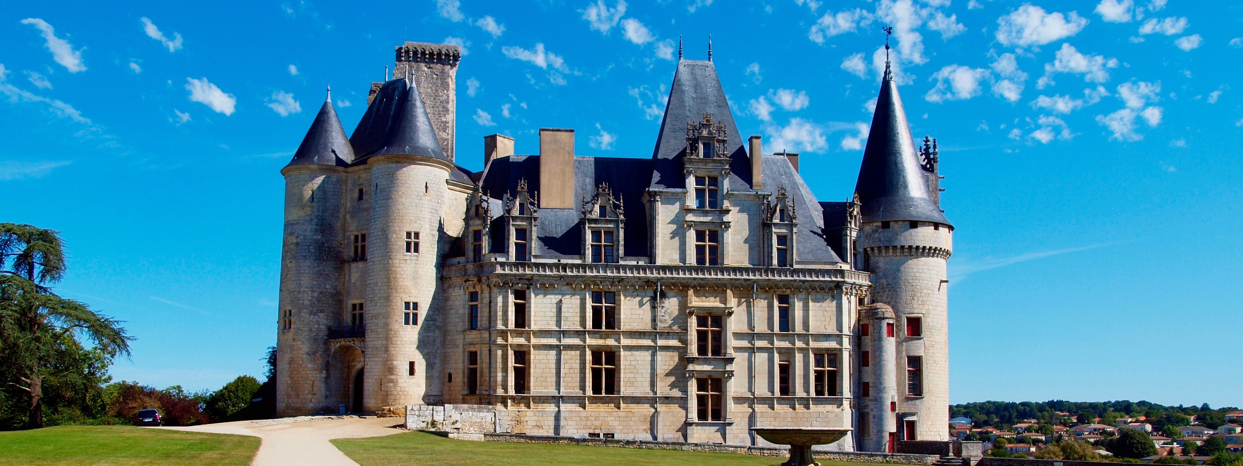 Depuis un chemin à côté d’un jardin d’herbe vert, vue sur l’aile sud du Château de la Rochefoucauld avec sa fontaine sous un ciel bleu.