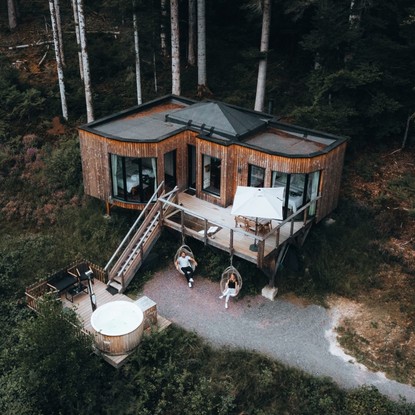 Maison moderne en bois avec une terrasse surélevée, un bain nordique et des fauteuils suspendus, située en pleine forêt.