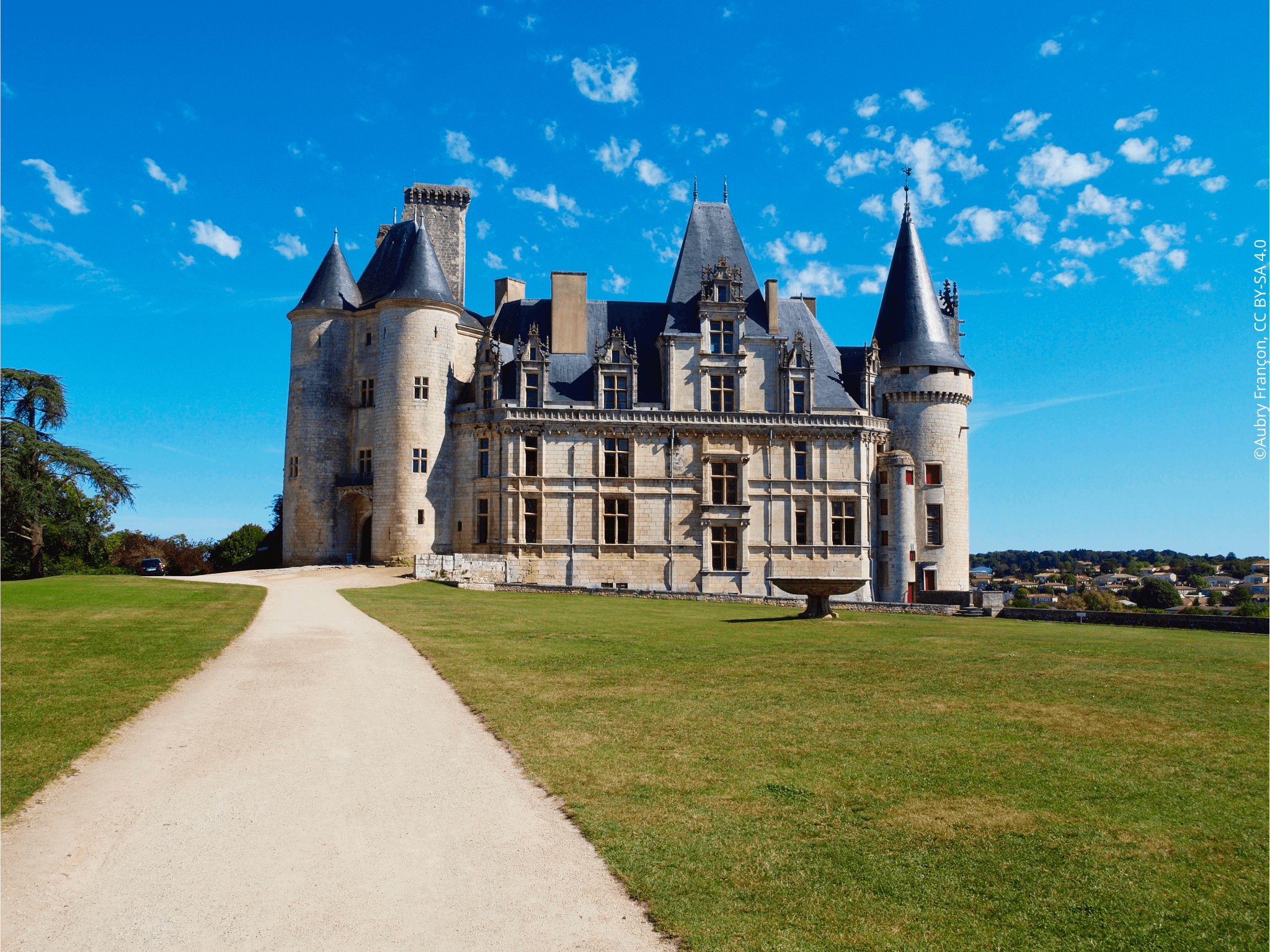 Depuis un chemin à côté d’un jardin d’herbe vert, vue sur l’aile sud du Château de la Rochefoucauld avec sa fontaine sous un ciel bleu.