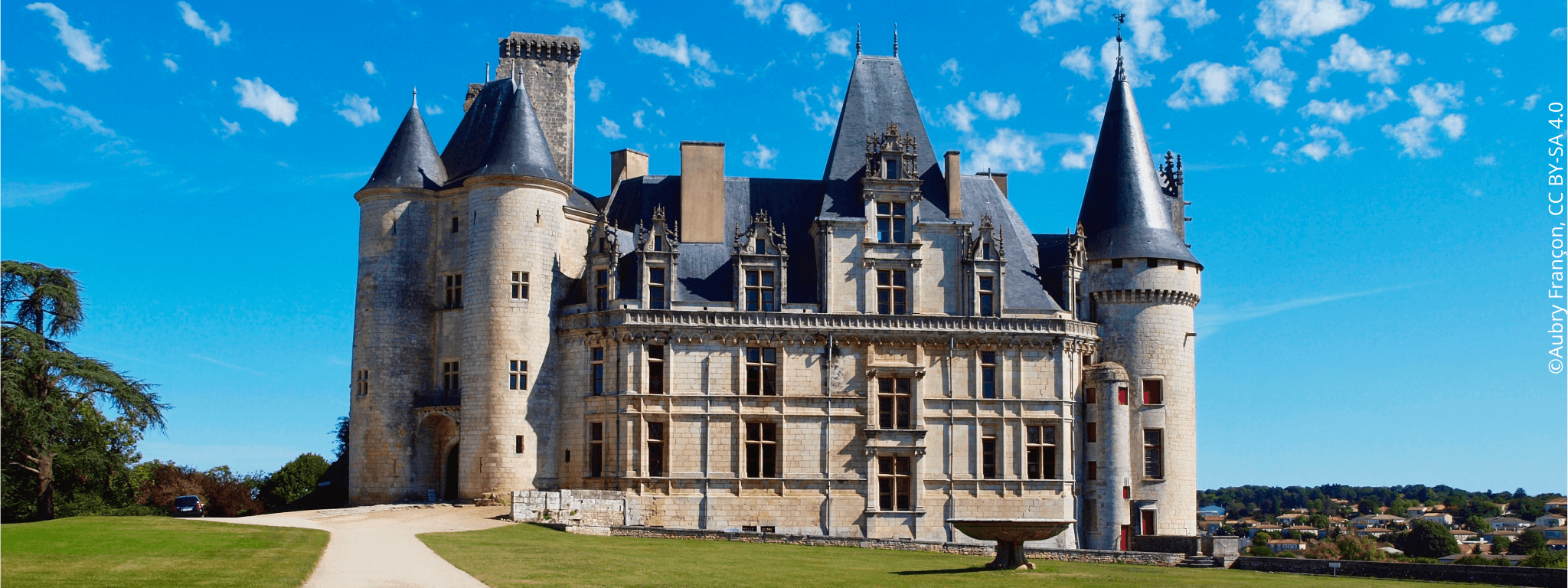 Depuis un chemin à côté d’un jardin d’herbe vert, vue sur l’aile sud du Château de la Rochefoucauld avec sa fontaine sous un ciel bleu.