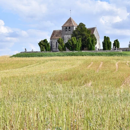 Église en pierre perchée sur une colline, entourée de champs dorés et d'arbres sous un ciel partiellement nuageux.