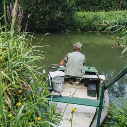 Homme pêchant au bord d'une rivière calme, entourée de végétation luxuriante et de fleurs sauvages.