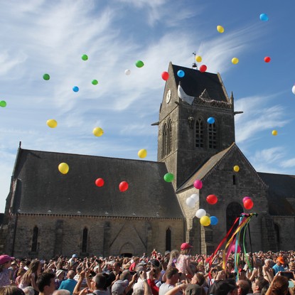 Une foule devant une eglise, soldat avec parachute blanc qui pend du clocher, ballons multicolores s'elevent dans le ciel bleu