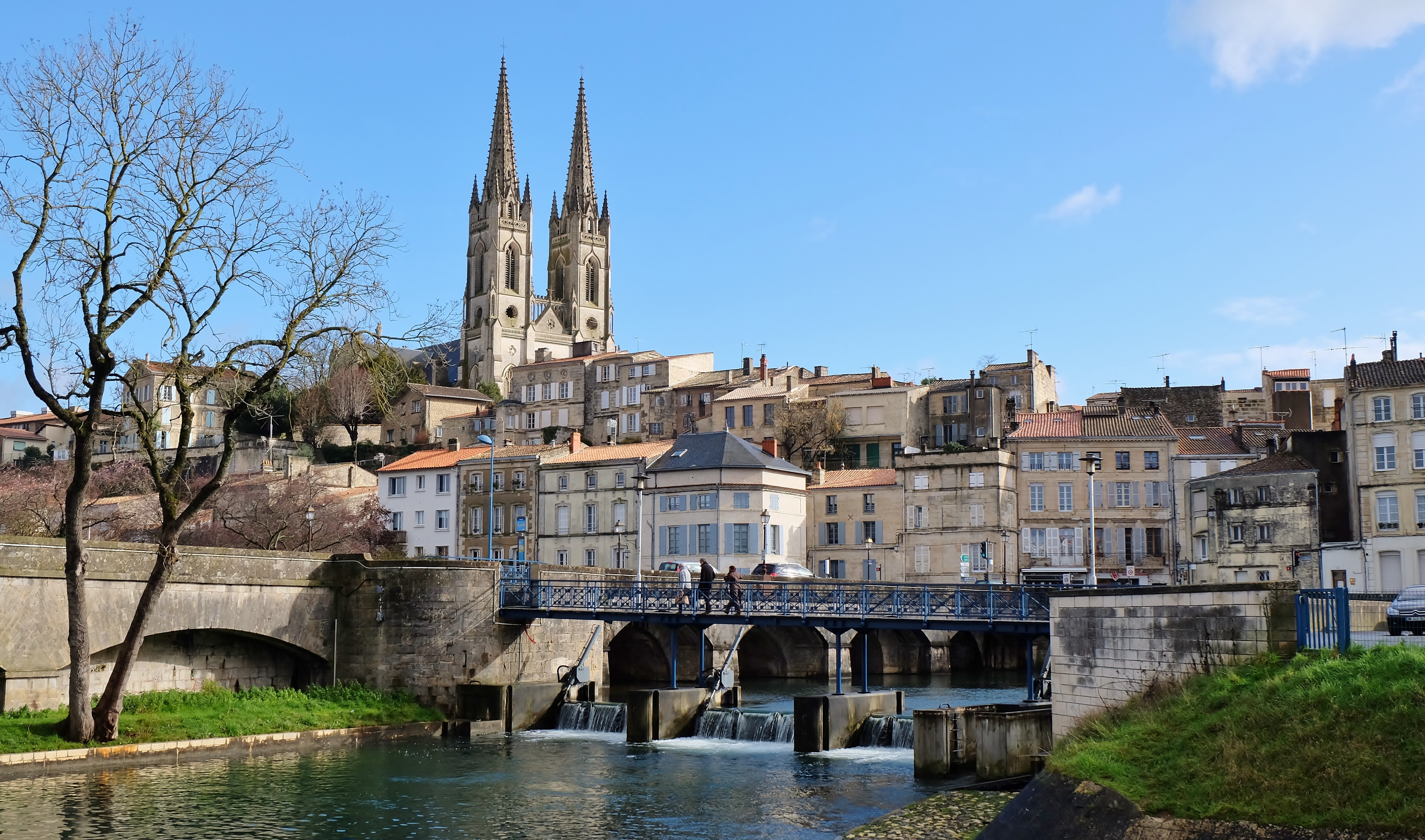 Pont traversant la Sèvre Niortaise menant à des habitations surélevées et l’église Saint André de Niort qui trône derrière.