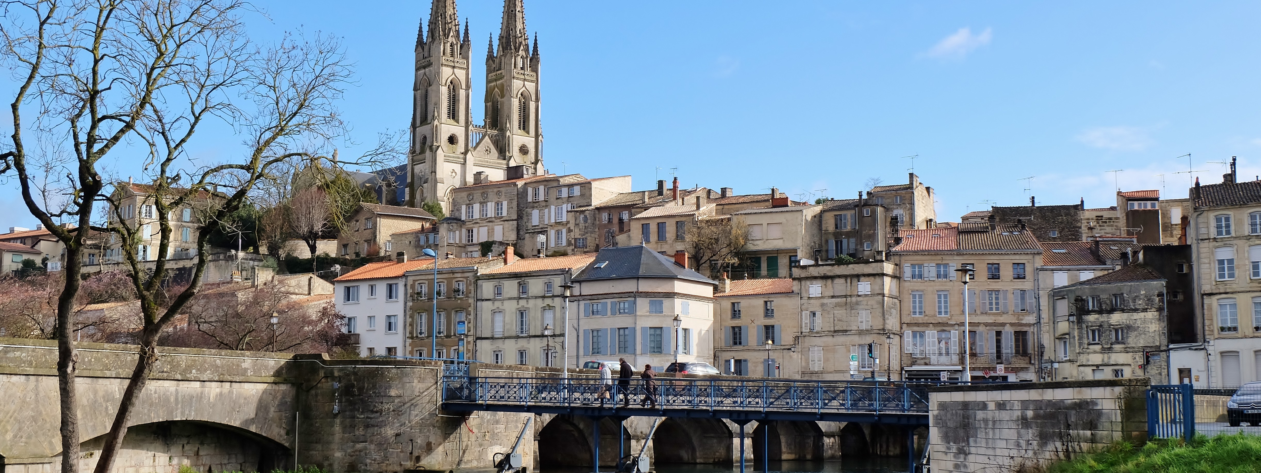 Pont traversant la Sèvre Niortaise menant à des habitations surélevées et l’église Saint André de Niort qui trône derrière.