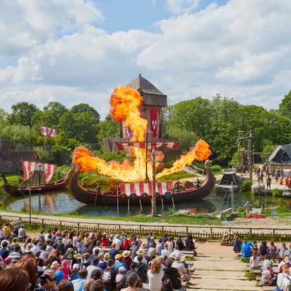 Spectacle du puy du Fou avec drakkar sur l’eau prenant feu et ce sous forme d'ancre, village gaulois pillé par les acteurs représentant des vikings et spectateurs en premier plan.