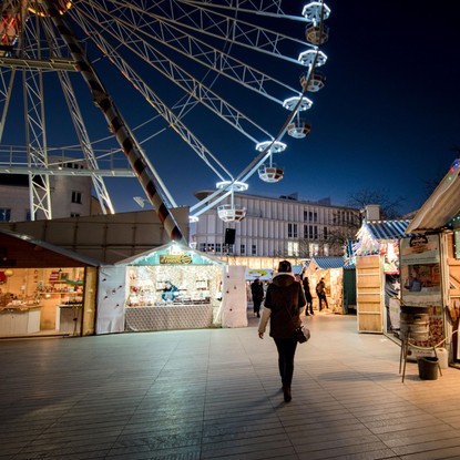 Marché de Noël la nuit, illuminée, avec une personne de dos qui marche, à droite un marchand d’épices et de pralines, avec d’autres chalets ouverts et éclairés ainsi que la grande roue illuminée à gauche