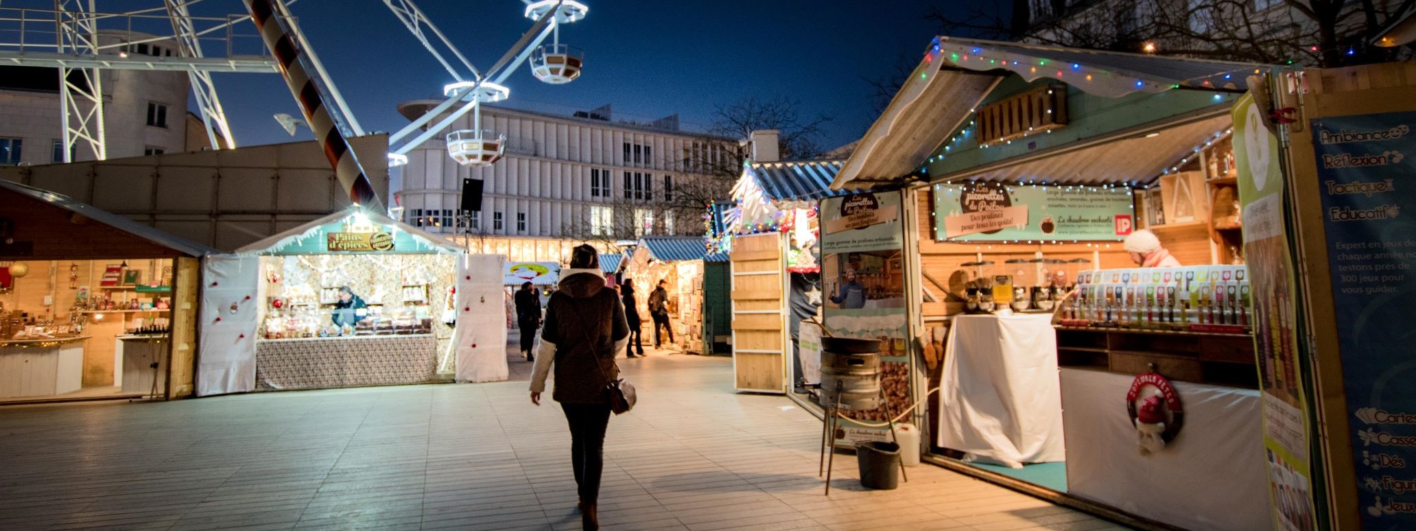 Marché de Noël la nuit, illuminée, avec une personne de dos qui marche, à droite un marchand d’épices et de pralines, avec d’autres chalets ouverts et éclairés ainsi que la grande roue illuminée à gauche