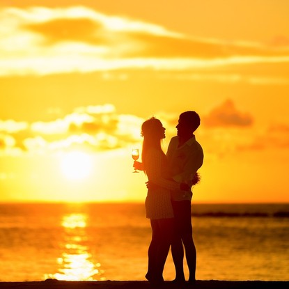 Un couple romantique face à un coucher de soleil doré au bord de l’océan, tenant un verre de vin.