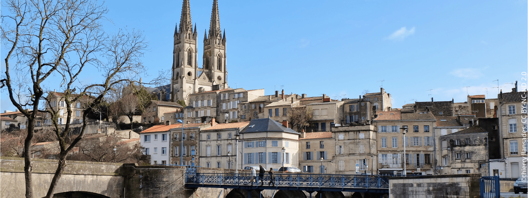 Pont traversant la Sèvre Niortaise menant à des habitations surélevées et l’église Saint André de Niort qui trône derrière.