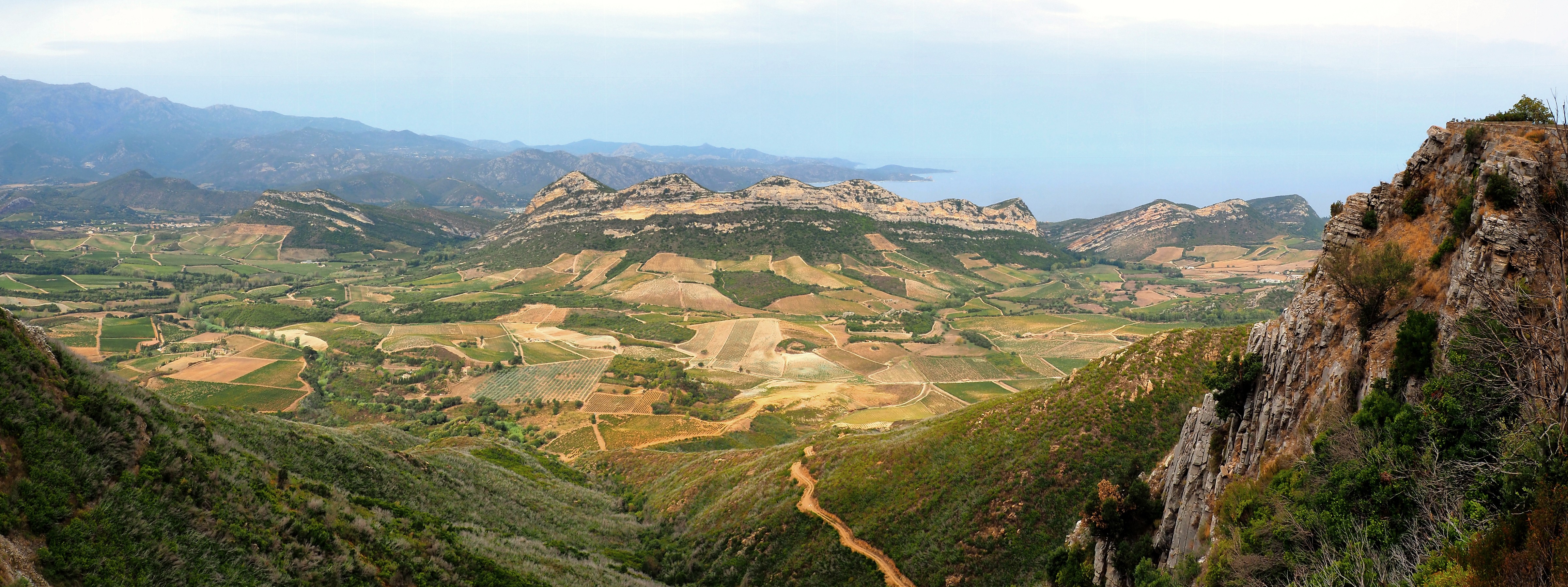 A panoramic view of Patrimonio between the mountains and the sea, with its mosaic of vineyards, lush green scrubland and winding paths. On the horizon, the Gulf of Saint-Florent completes this emblematic landscape of the Nebbiu.