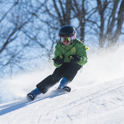 Jeune garçon en combinaison verte dévalant une piste de ski avec un casque et des lunettes, éclats de neige en arrière-plan.