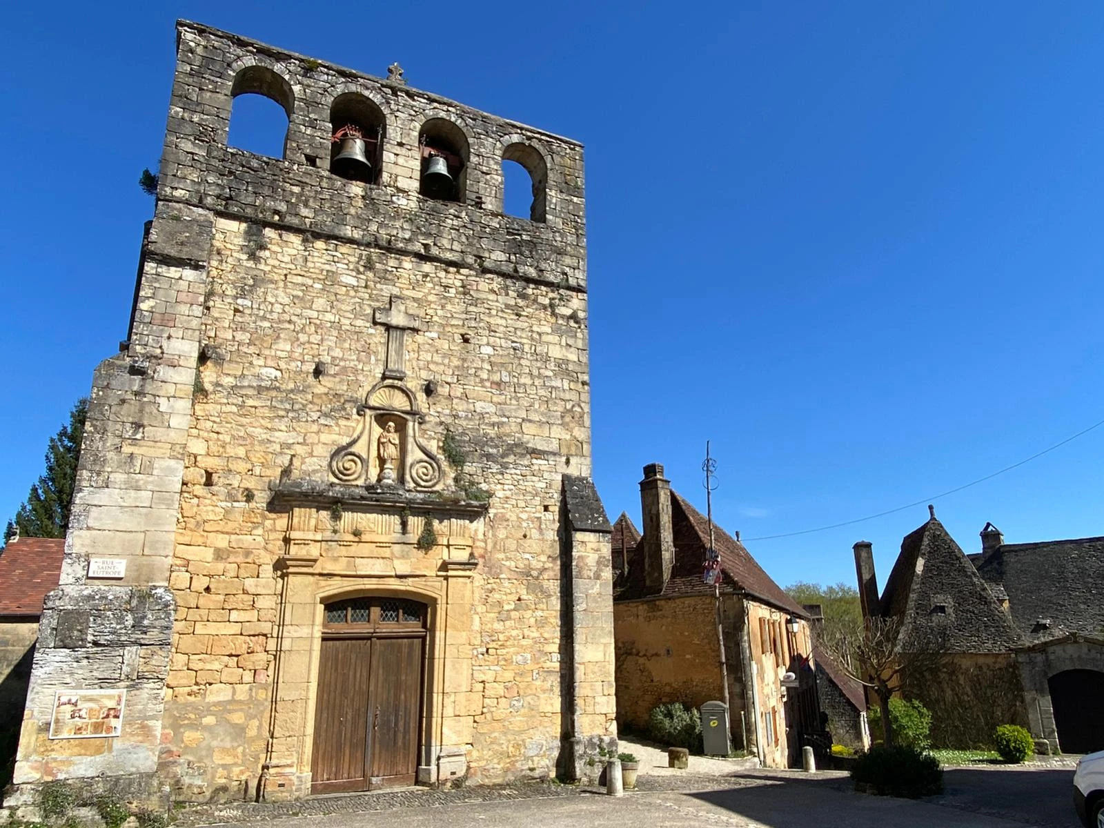Façade de l’église Saint-Eutrope de Meyrals en Dordogne, typique de l’architecture du Périgord Noir, avec son clocher-mur et ses pierres blondes, sous un ciel bleu éclatant. Le charme authentique d’un village périgourdin.