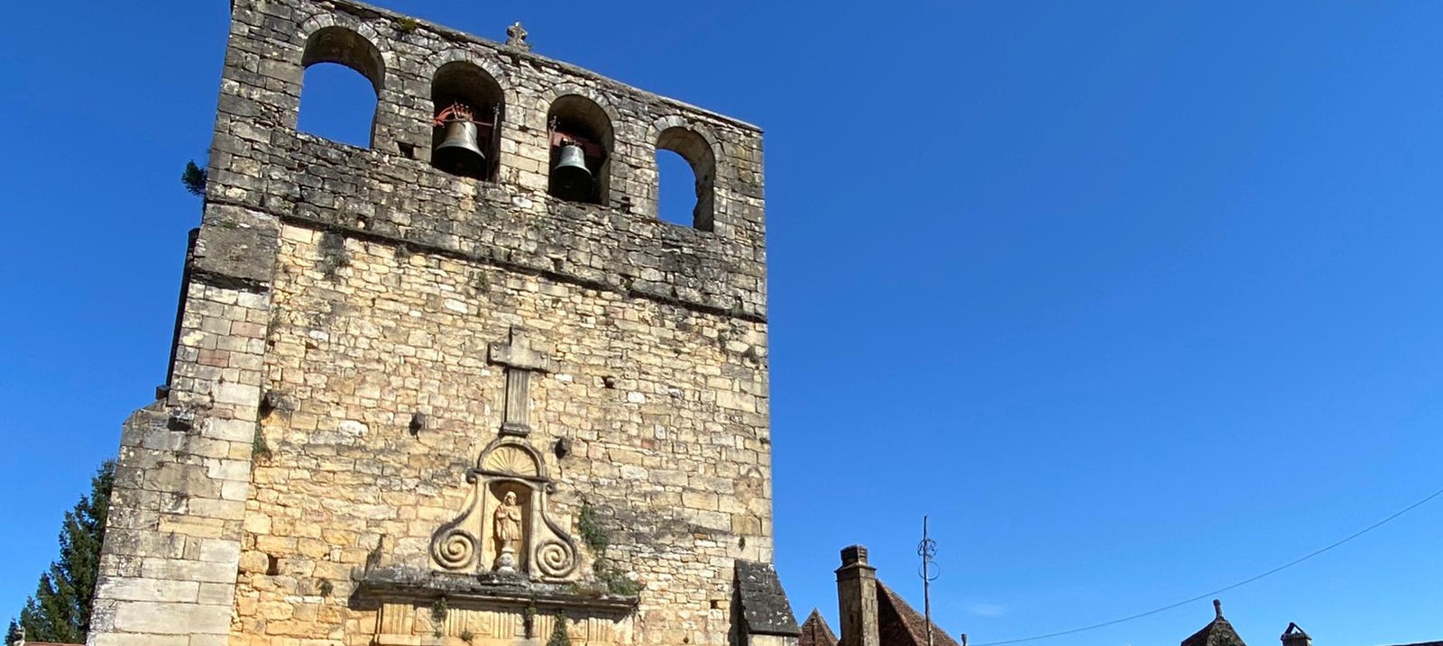 Façade de l’église Saint-Eutrope de Meyrals en Dordogne, typique de l’architecture du Périgord Noir, avec son clocher-mur et ses pierres blondes, sous un ciel bleu éclatant. Le charme authentique d’un village périgourdin.