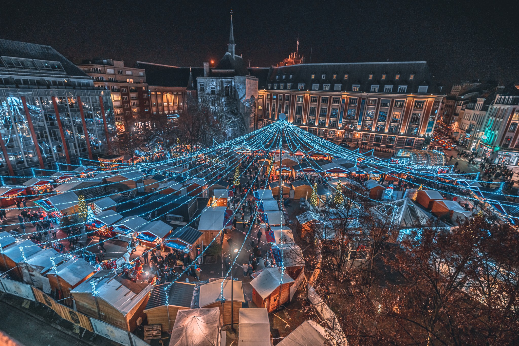 Vue aérienne d'un marché de Noël illuminé avec des chalets en bois, des guirlandes lumineuses suspendues en forme de toile au-dessus des stands, et des bâtiments décorés entourant la place. Les visiteurs se promènent parmi les stands sous les décorations festives.