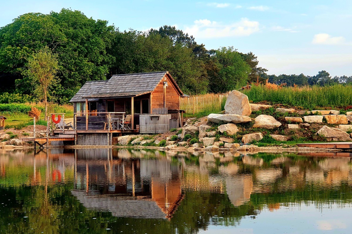 Cabane en bois au bord d'un étang paisible, avec une terrasse et des chaises, entourée de verdure.