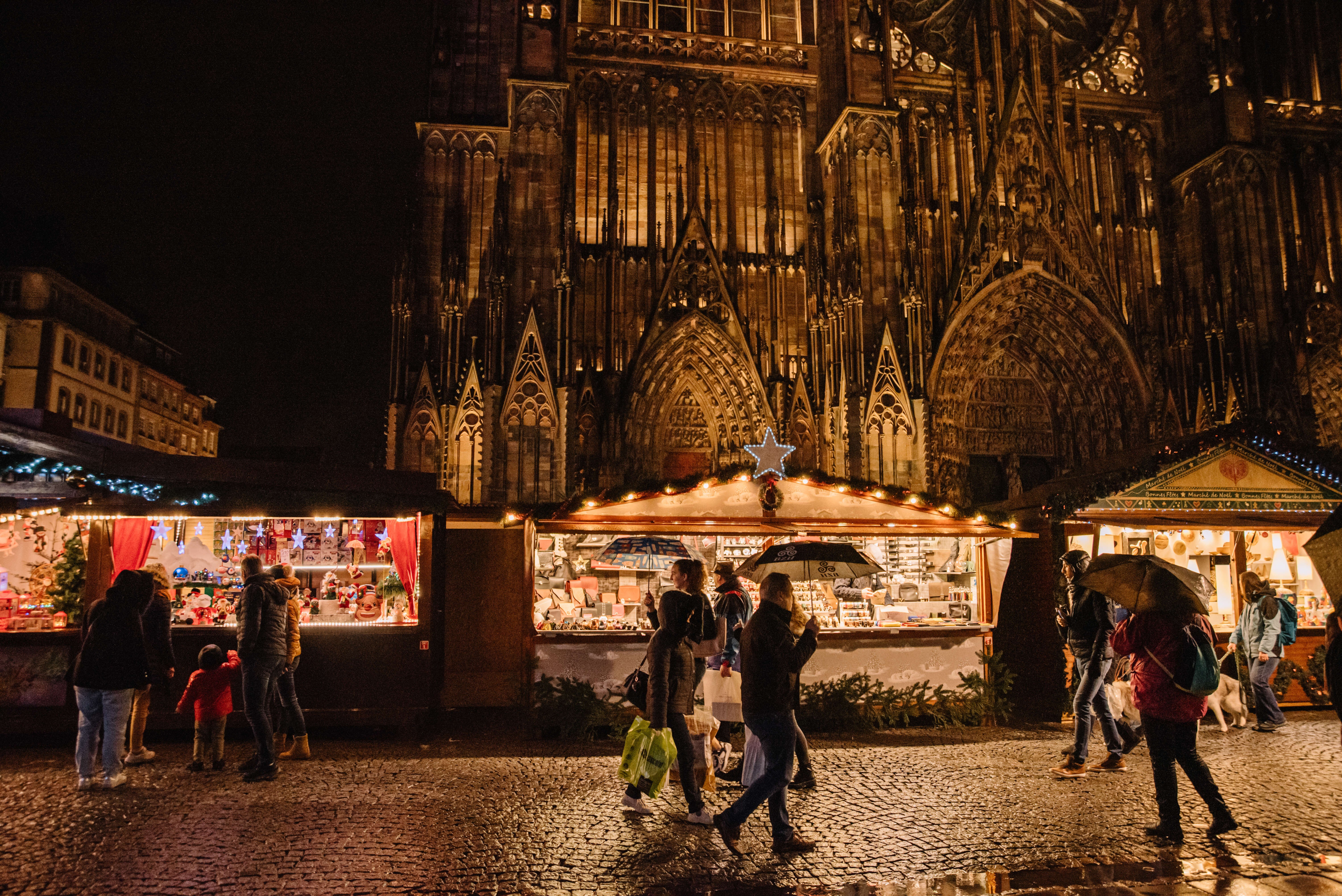 Marché de Noël de Strasbourg devant la cathédrale illuminée, avec des stands décorés proposant des produits artisanaux et des visiteurs déambulant sous la pluie avec des parapluies, créant une ambiance festive et chaleureuse.