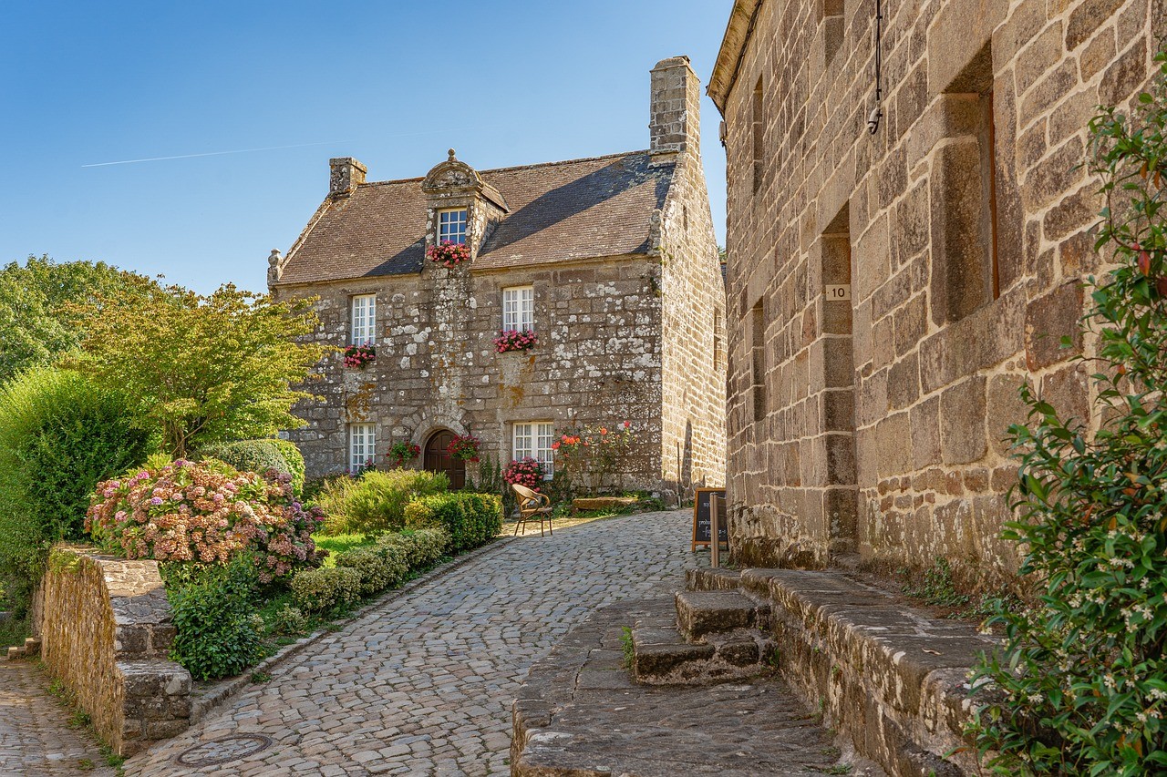 Village pittoresque avec des maisons en pierre, un chemin pavé et des fleurs colorées sous un ciel bleu.