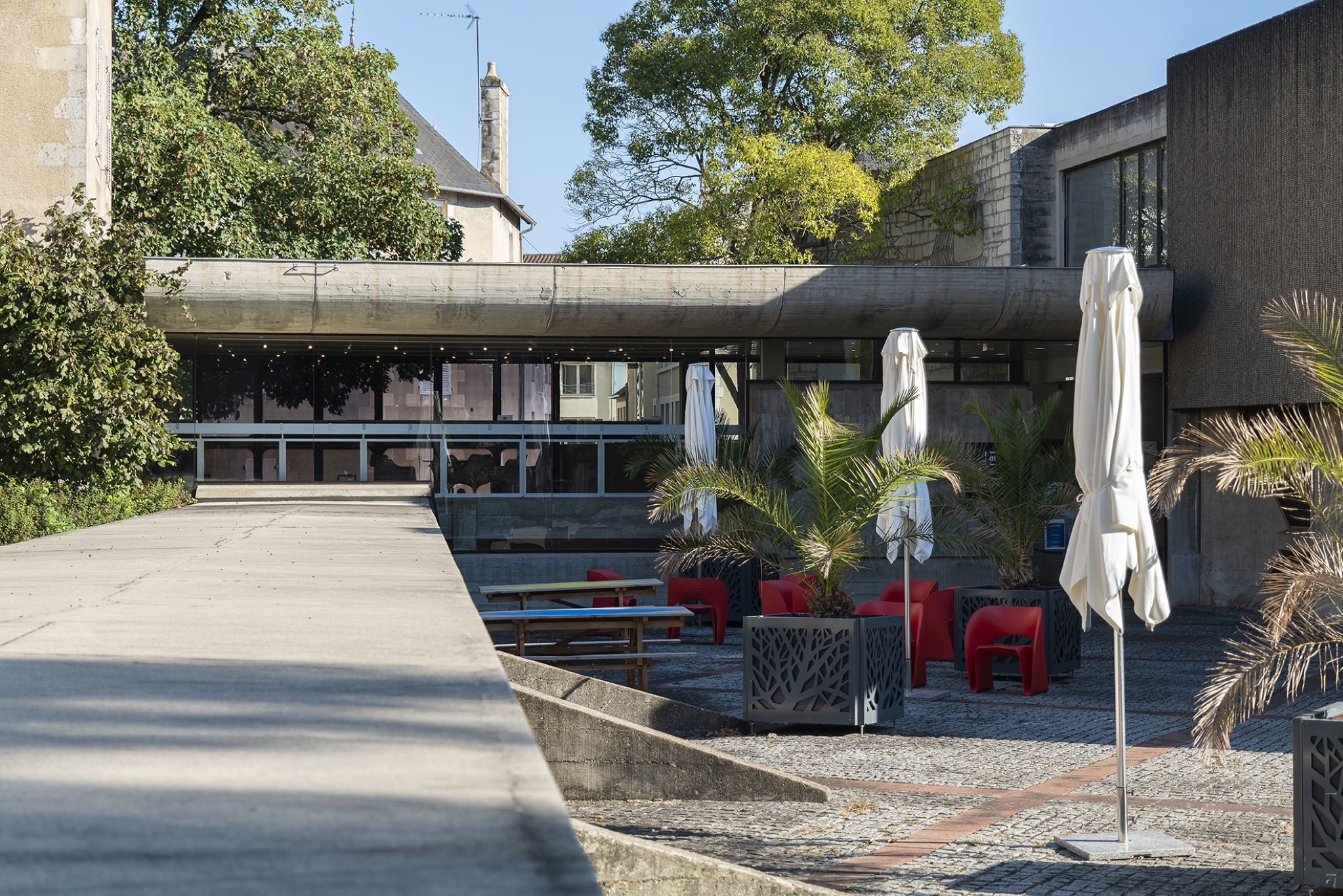 Entrée du Musée Ste Croix à Poitiers avec trois parasols fermés, 2 palmiers en pots et des fauteuils rouges avec des tables et des bancs sur l’espace repos, avec une coursive vitrée dans le fond qui passe d’un bâtiment à un autre