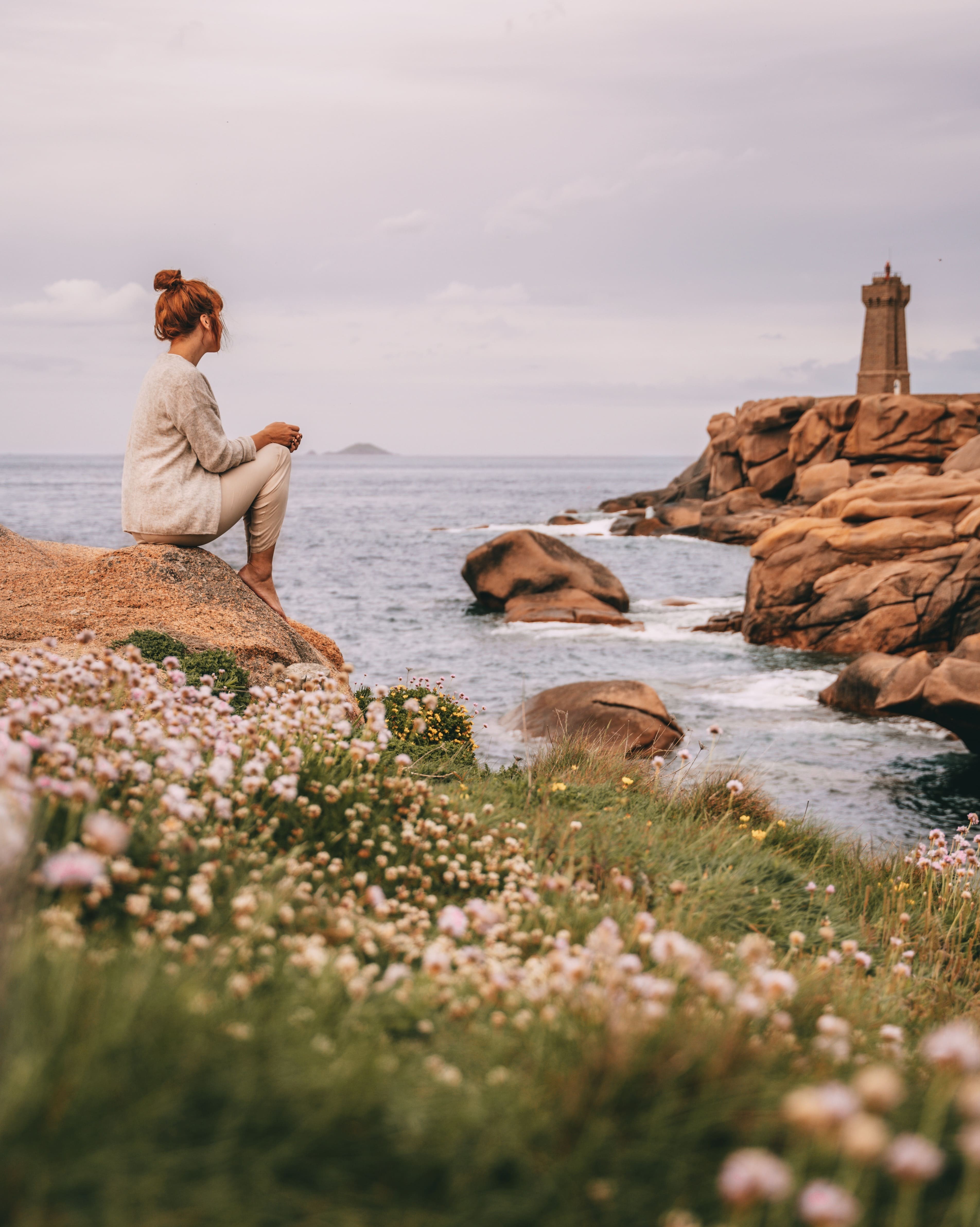 Jeune femme rousse assise sur rocher de la côte de Granit Rose regardant le phare de Ploumanach