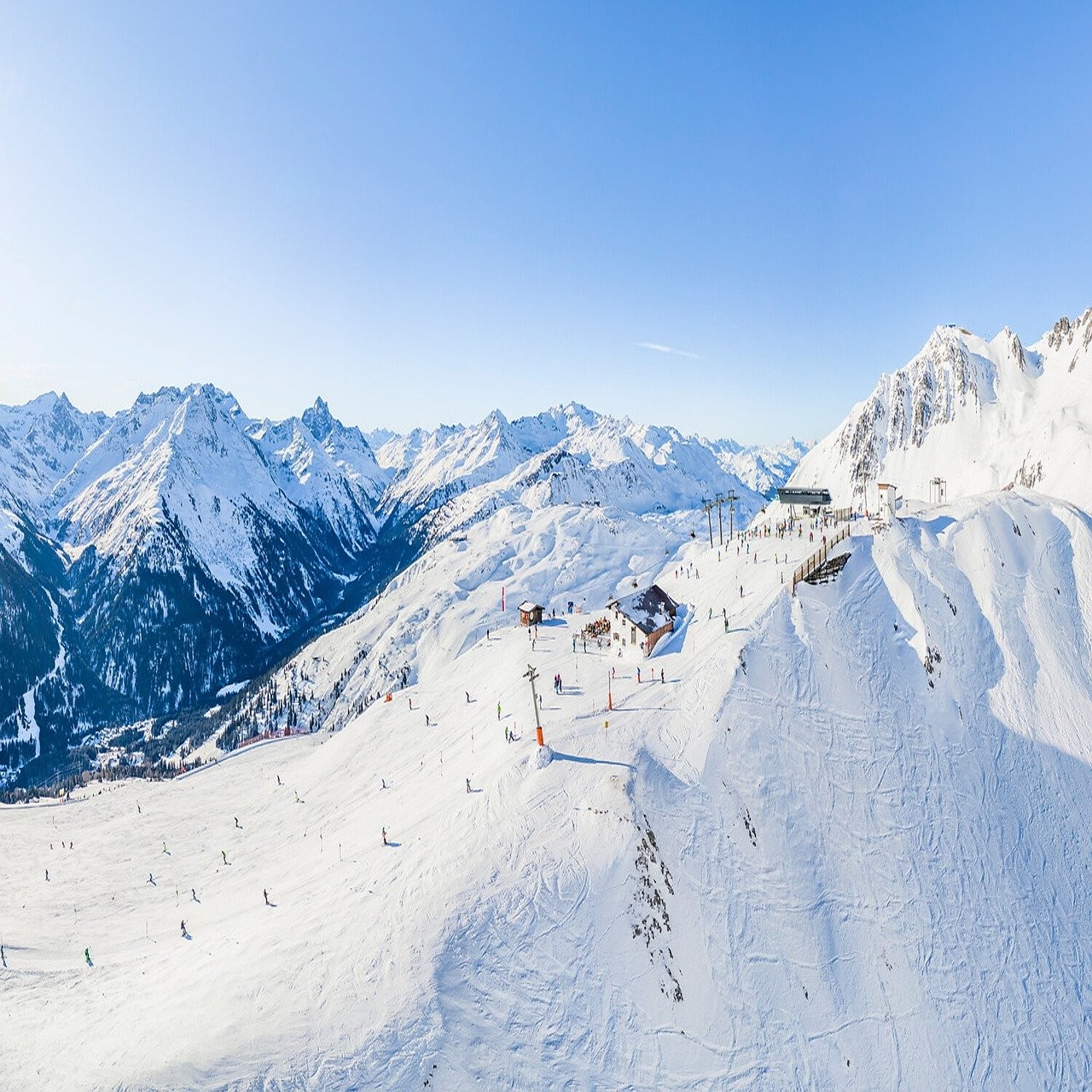 Panorama aérien d'une station de ski avec des pistes enneigées et des montagnes en arrière-plan, sous un ciel bleu clair.