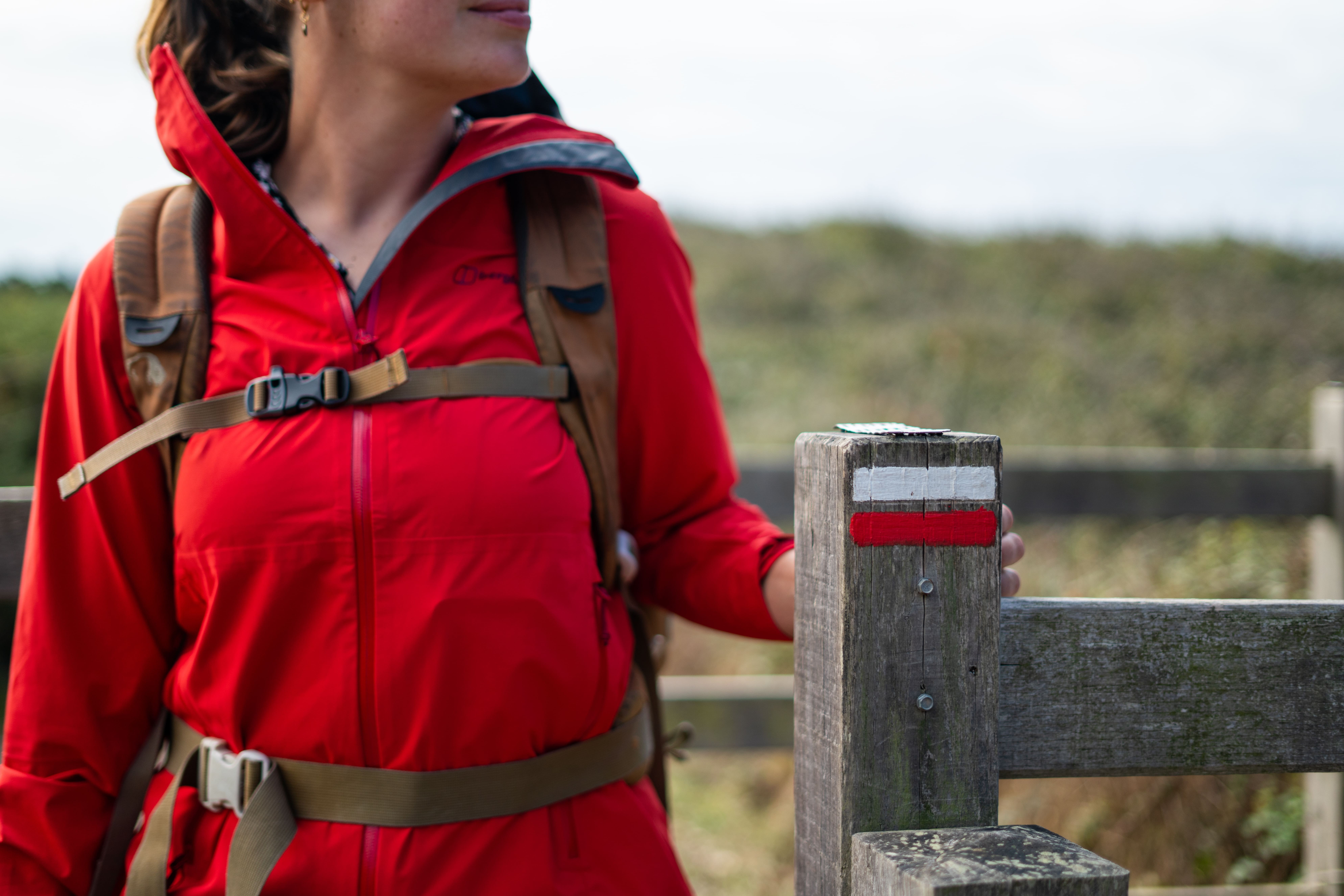 Jeune femme avec manteau rouge et sac à dos sur le sentier des douaniers avec balise du GR34 sur poteau bois
