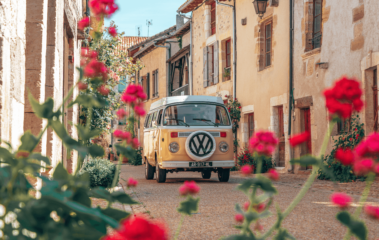 Un minibus VW roulant dans une rue étroite bordée de fleurs rouges à Saint-Jean-de-Côle
