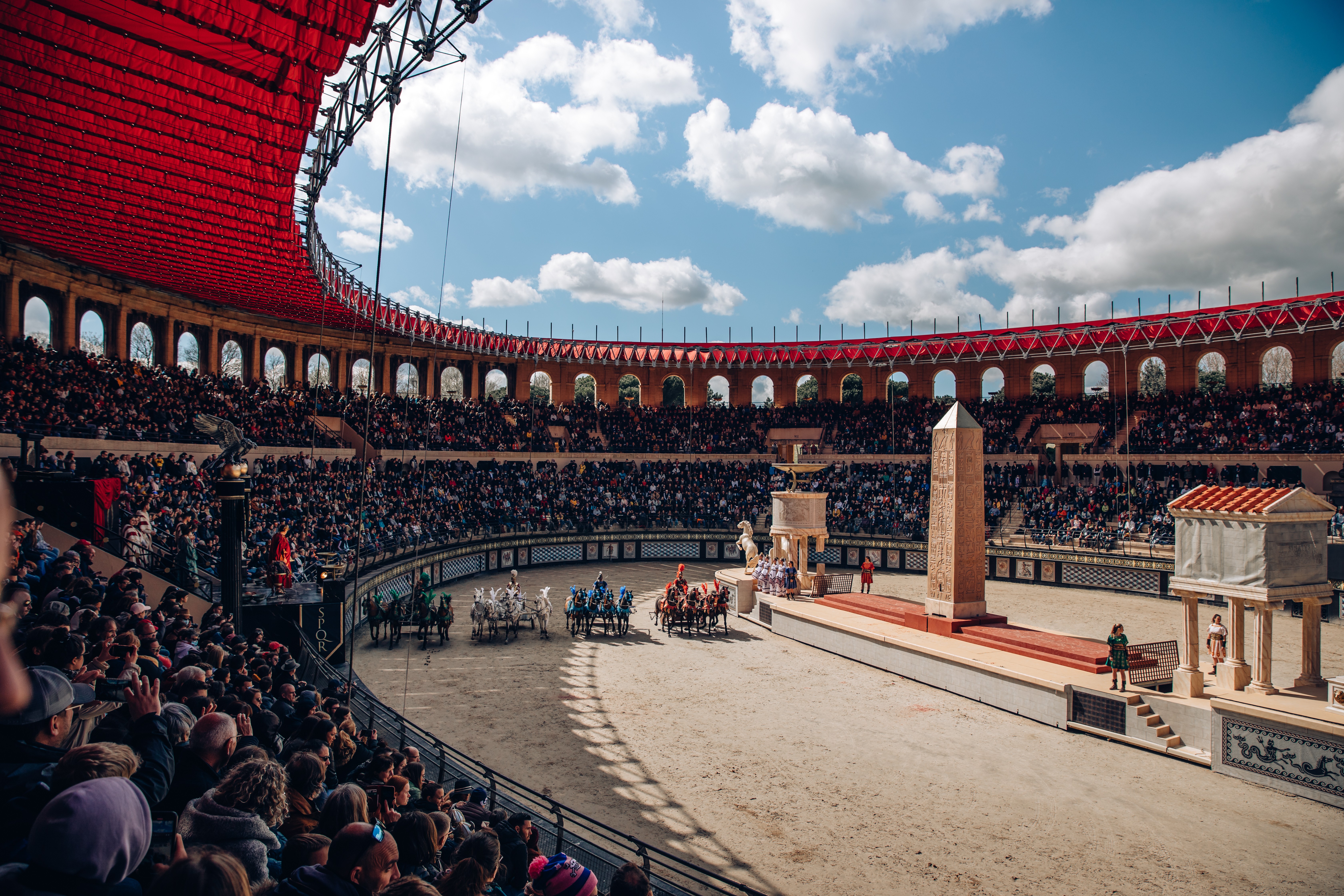 Spectacle du Puy du Fou dans une arène couverte d’une toile rouge avec 4 équipes de chevaux menés par des gladiateurs et des spectateurs autour dans les gradins