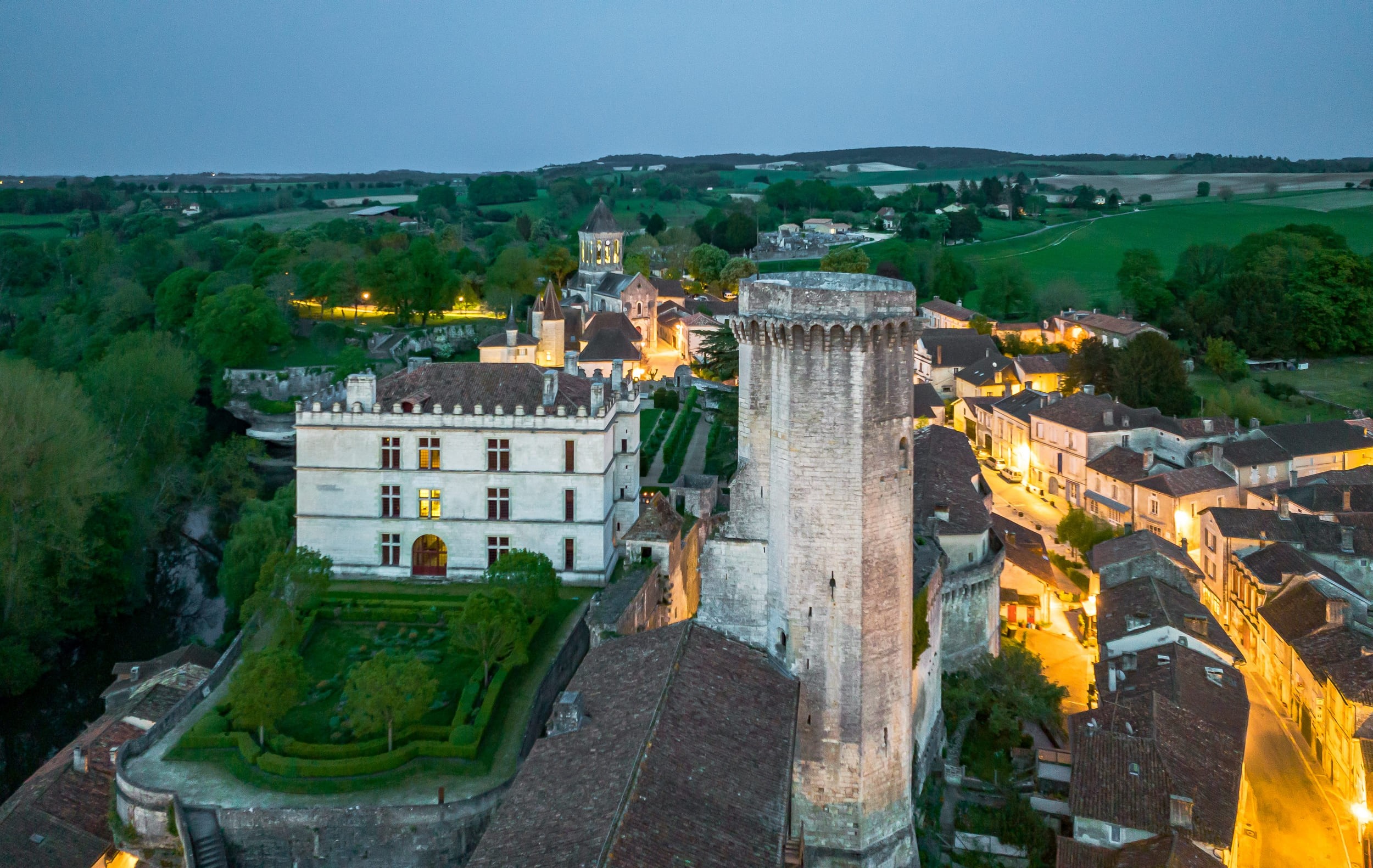 Vue aérienne d'une ville médiévale au crépuscule avec le château de Bourdeilles illuminé.