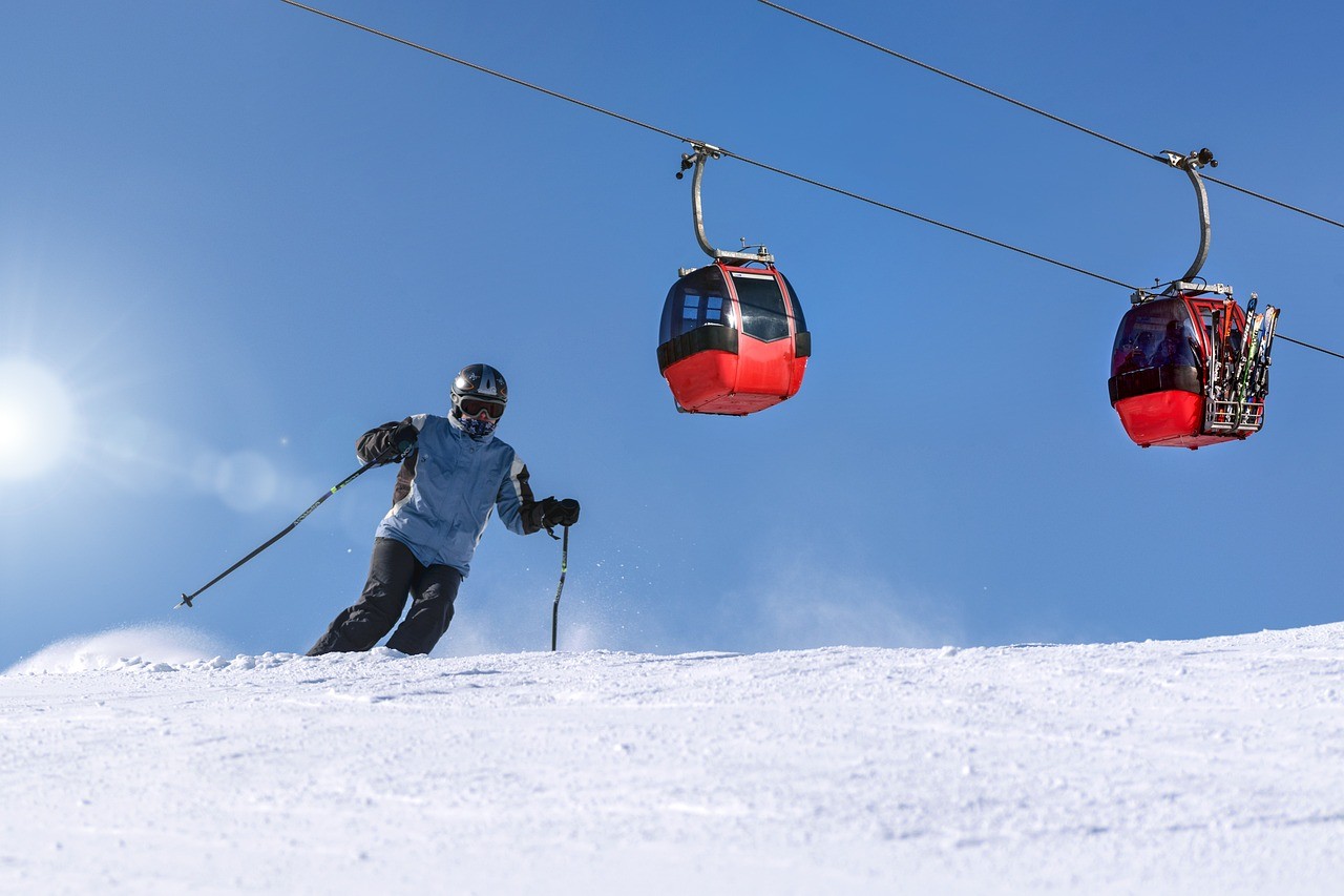 Skieur dévalant une piste enneigée sous un ciel bleu, avec deux télécabines rouges en arrière-plan.