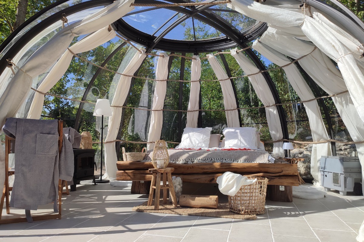 Dôme transparent aménagé avec un lit confortable et une décoration chaleureuse, entouré de nature verdoyante