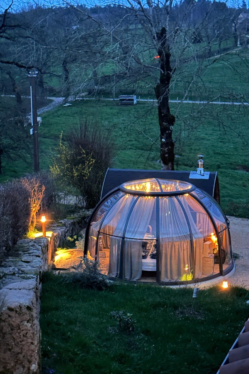 Une bulle d’hébergement lumineuse et chaleureuse dans un cadre rural au crépuscule, entourée de verdure et éclairée par des lampes extérieures.