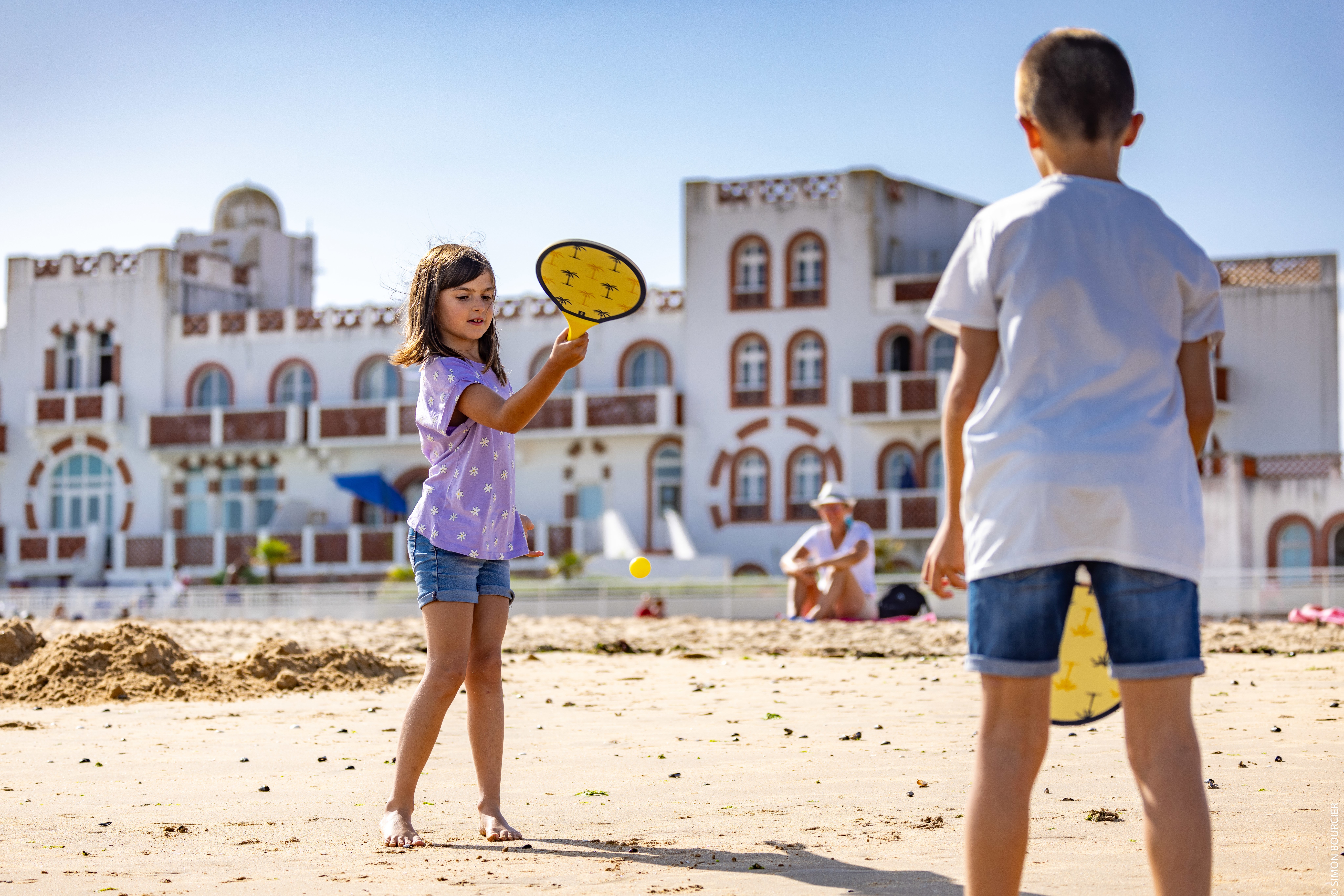 Deux enfants jouent aux raquettes sur la plage centrale devant l’immeuble au style oriental