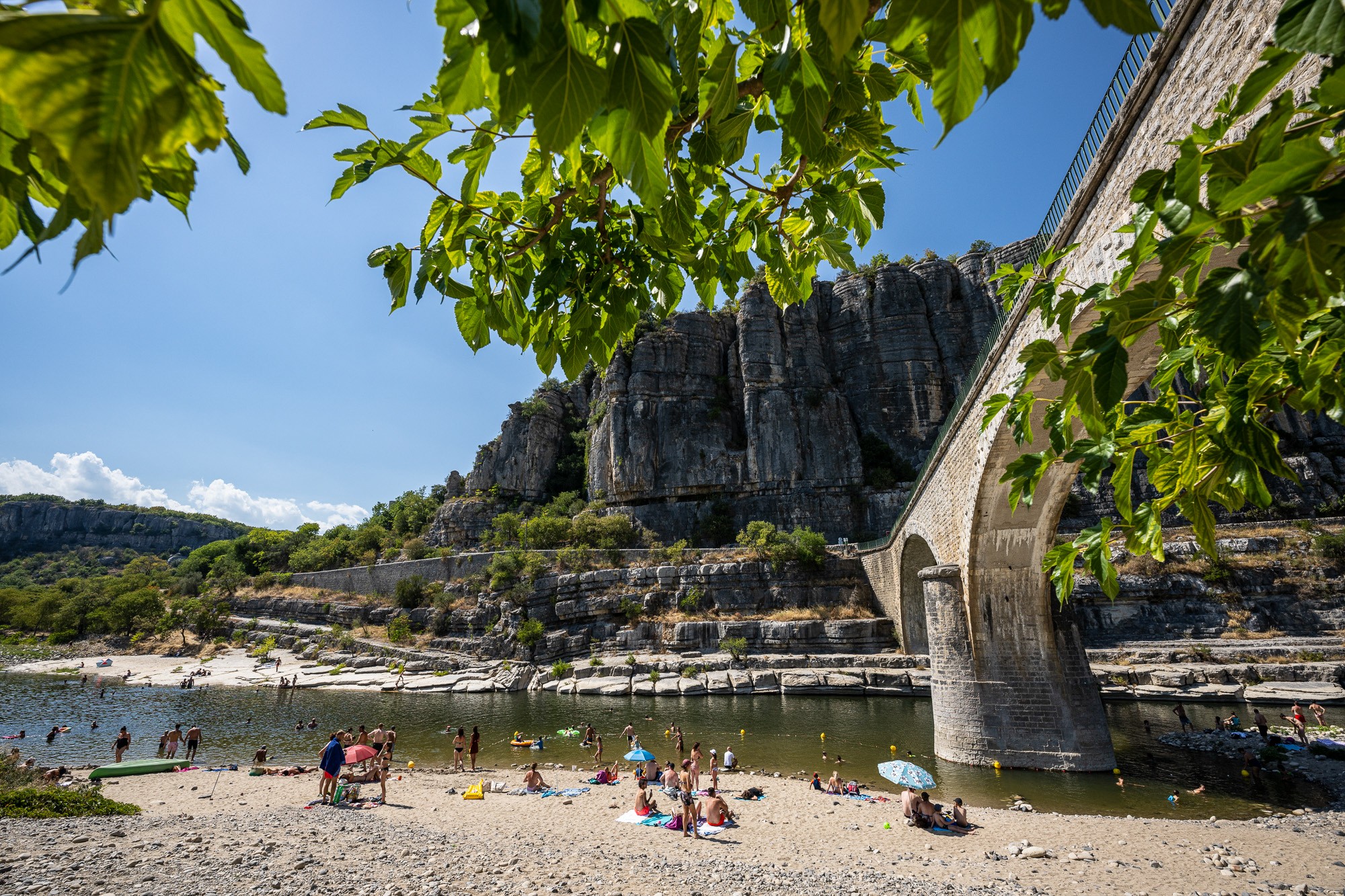 Village de caractère de Balazuc - vue depuis la plage avec des touristes qui se baignent dans la rivière Ardèche et le pont