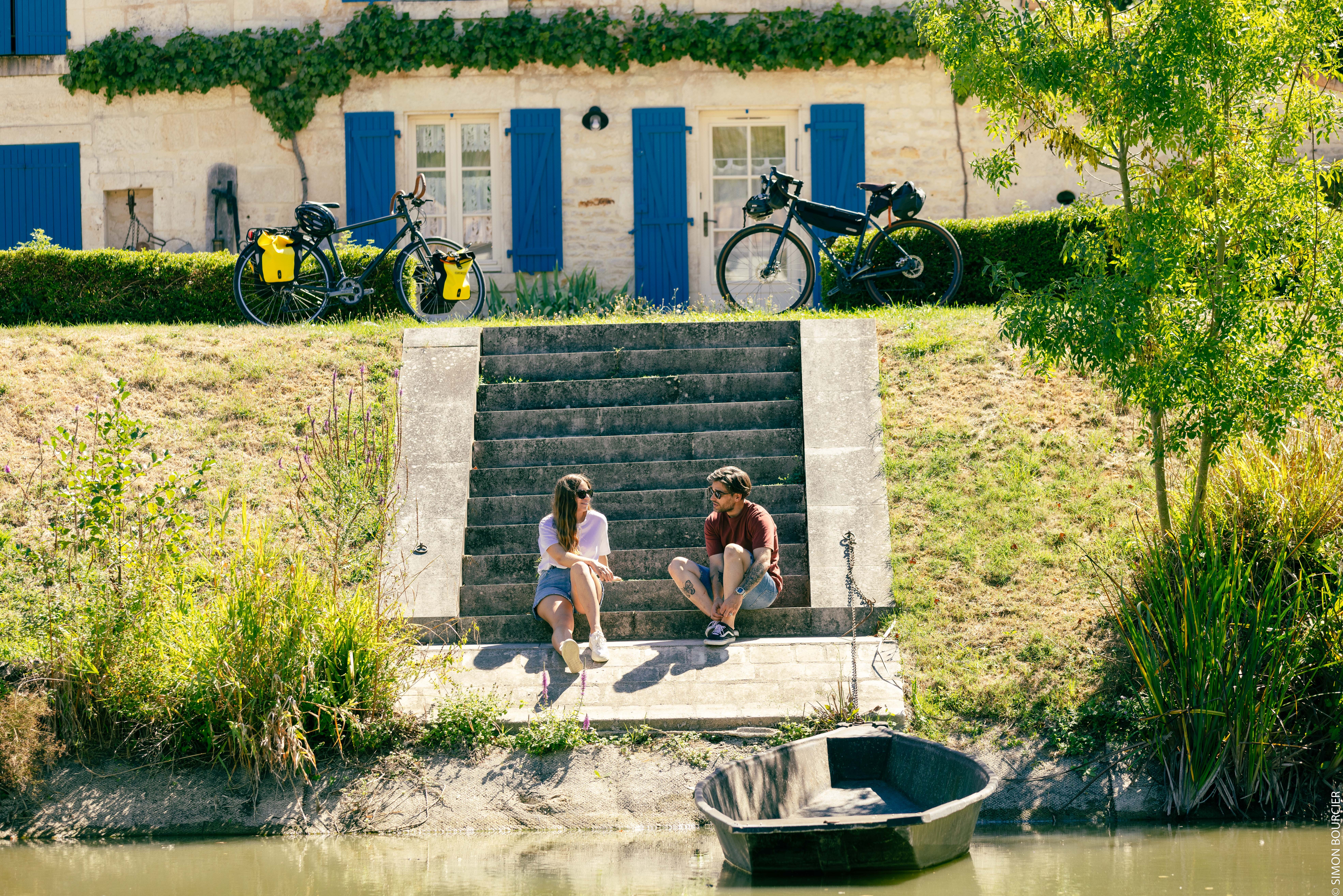 Un homme et une femme ayant stationné leur vélos le long d’une longère maraichine sont assis sur des marches en bordure de marais Poitevin avec une barque à leurs pieds.
