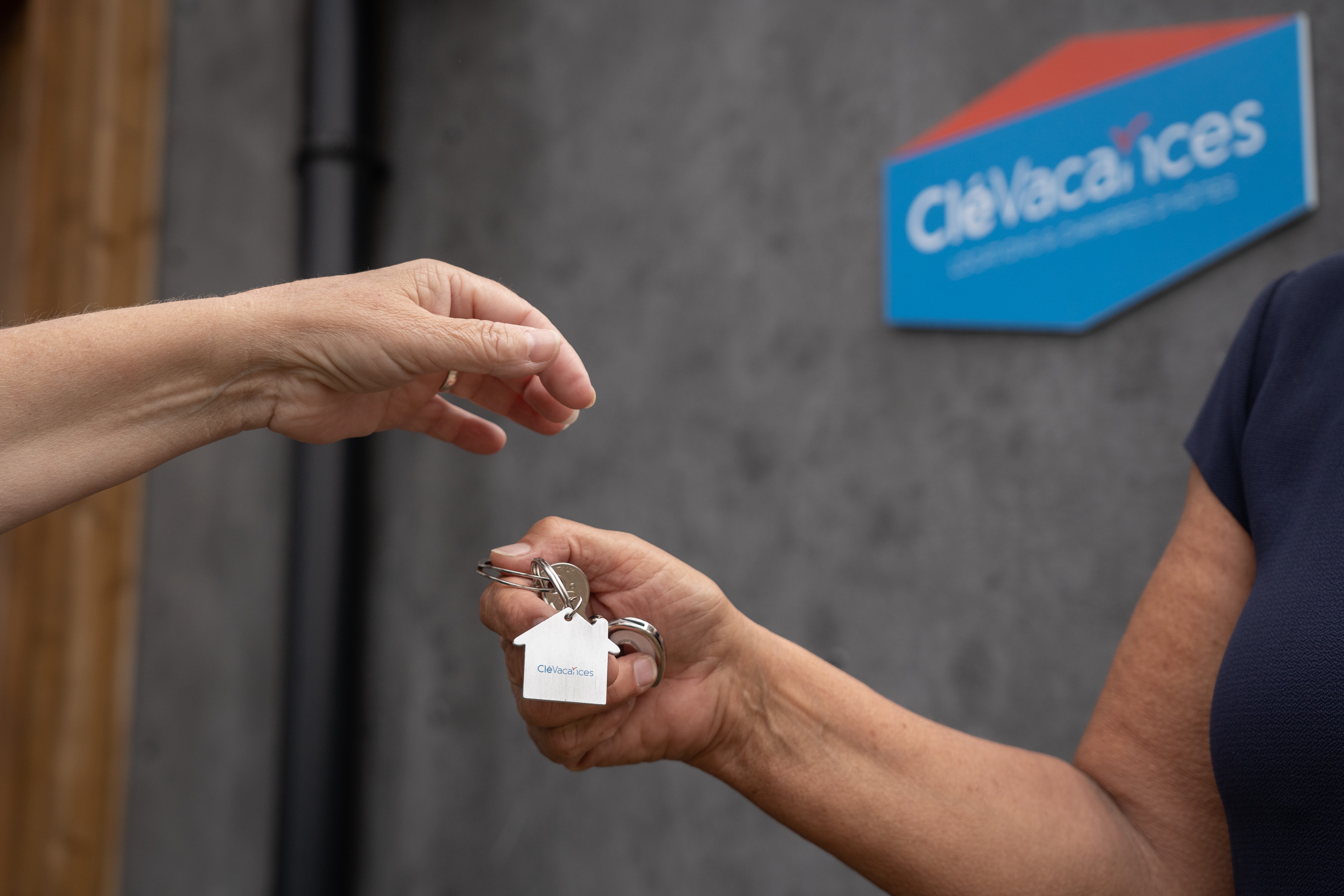 Photo cadrée sur les mains de deux personnes se donnant un trousseau de clés. Un panneau Clévacances est visible en arrière-plan.