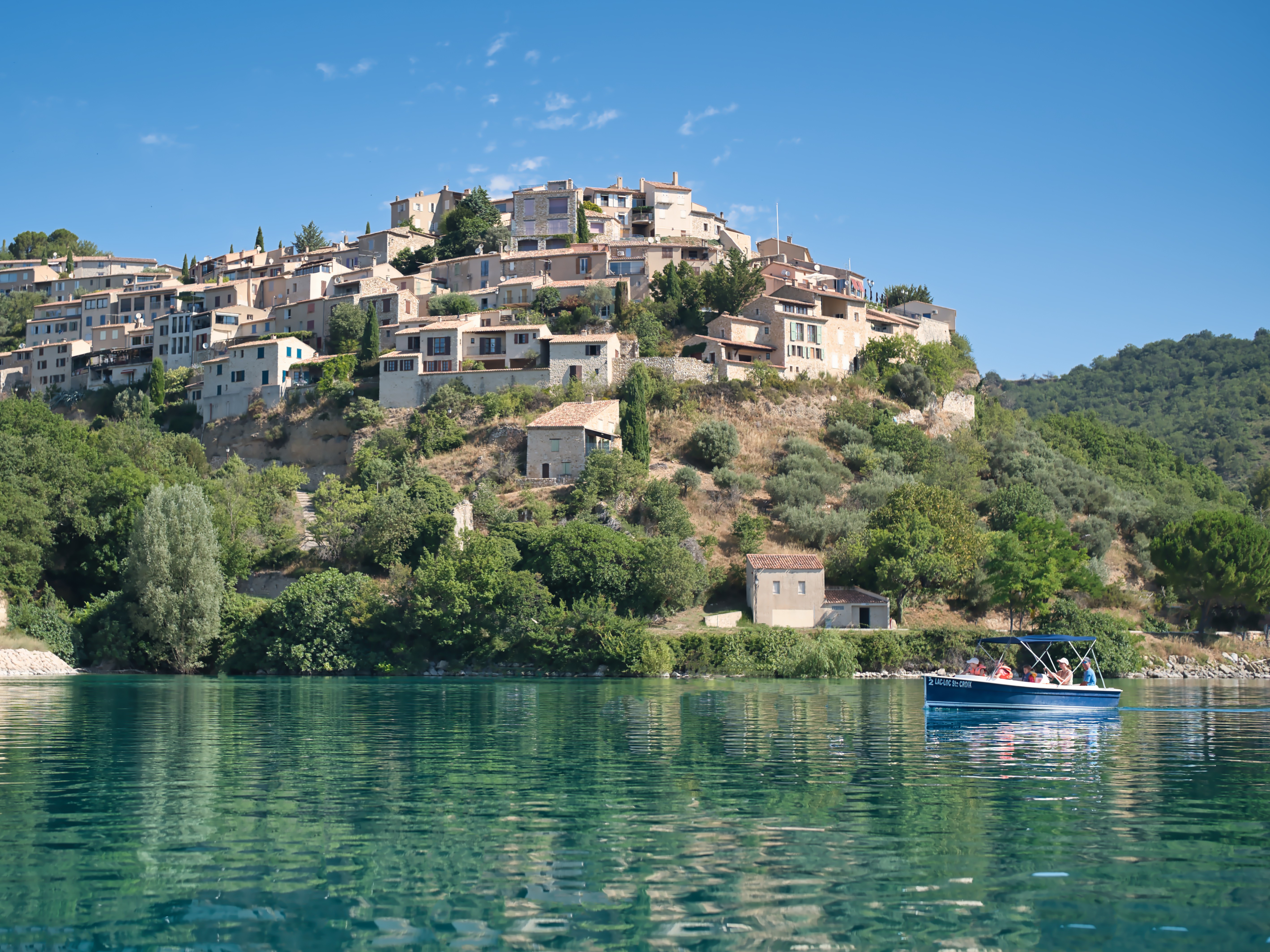 Le village perché de Sainte-Croix-du-Verdon vu depuis les eaux calmes du Lac de Sainte-Croix avec un bateau de tourisme au premier plan