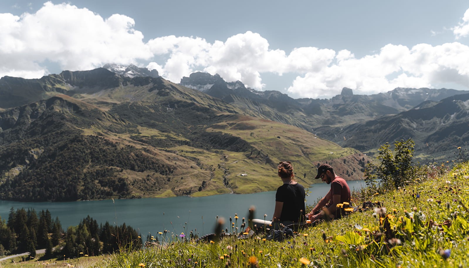 Deux personnes assises sur une prairie alpine, profitant d'une vue panoramique sur un lac de montagne et des sommets majestueux sous un ciel partiellement nuageux