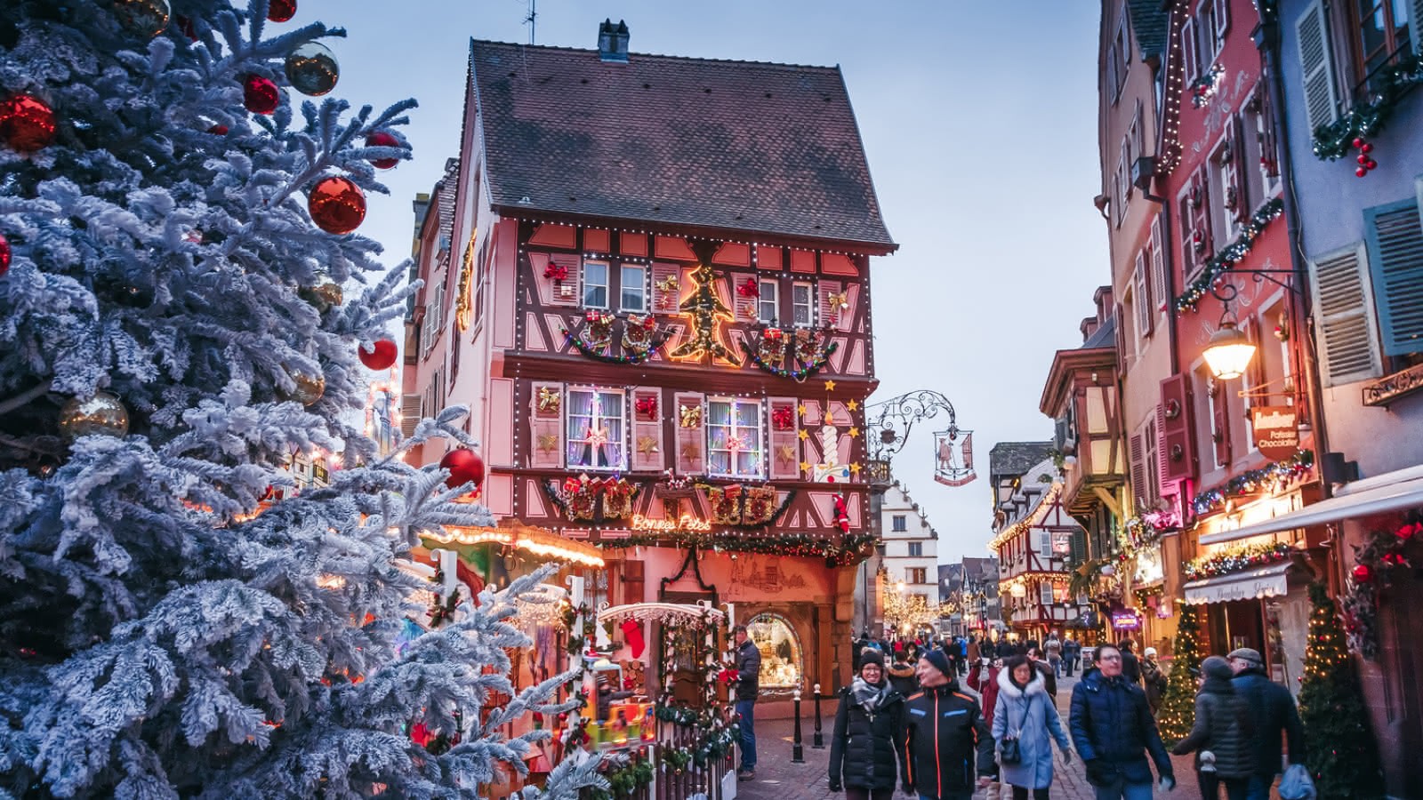 Marché de Noël à Colmar, avec des maisons à colombages décorées, un grand sapin enneigé orné de boules rouges et dorées, et des visiteurs se promenant dans les rues illuminées.