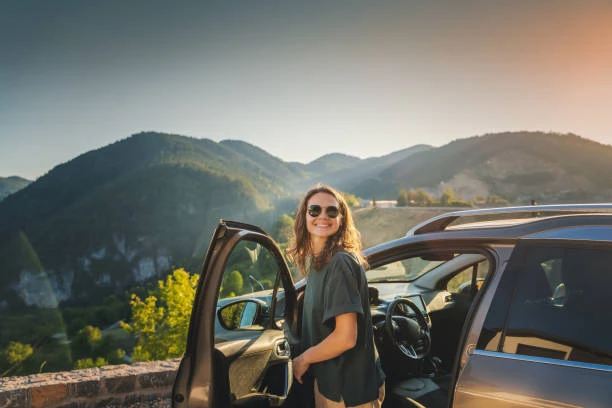 Femme souriante près de sa voiture avec une vue panoramique sur des collines verdoyantes.