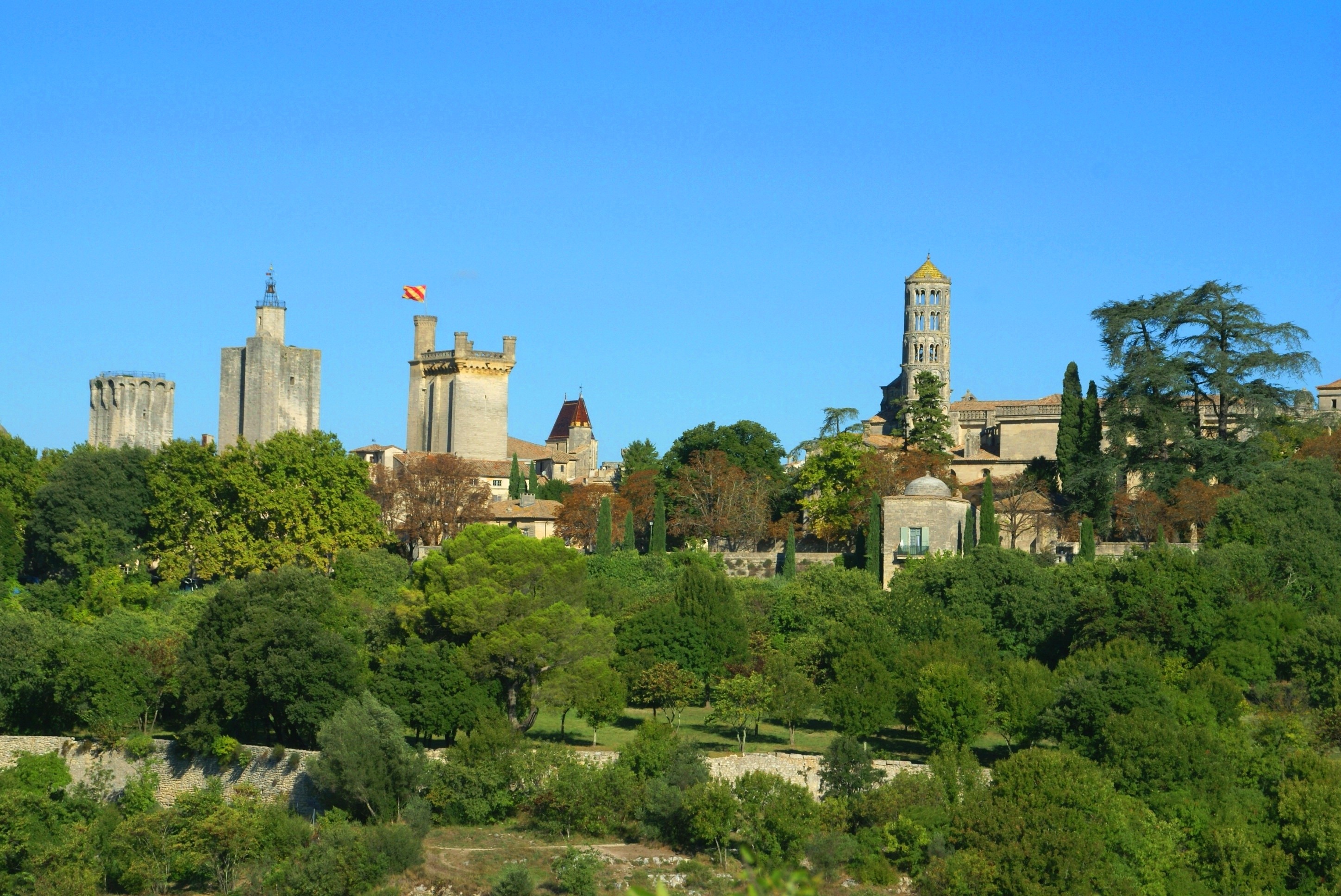 Vue panoramique de la cité ducale d’Uzès. Végétation verdoyante en premier plan, les tours de la cité se détachent sur le ciel bleu