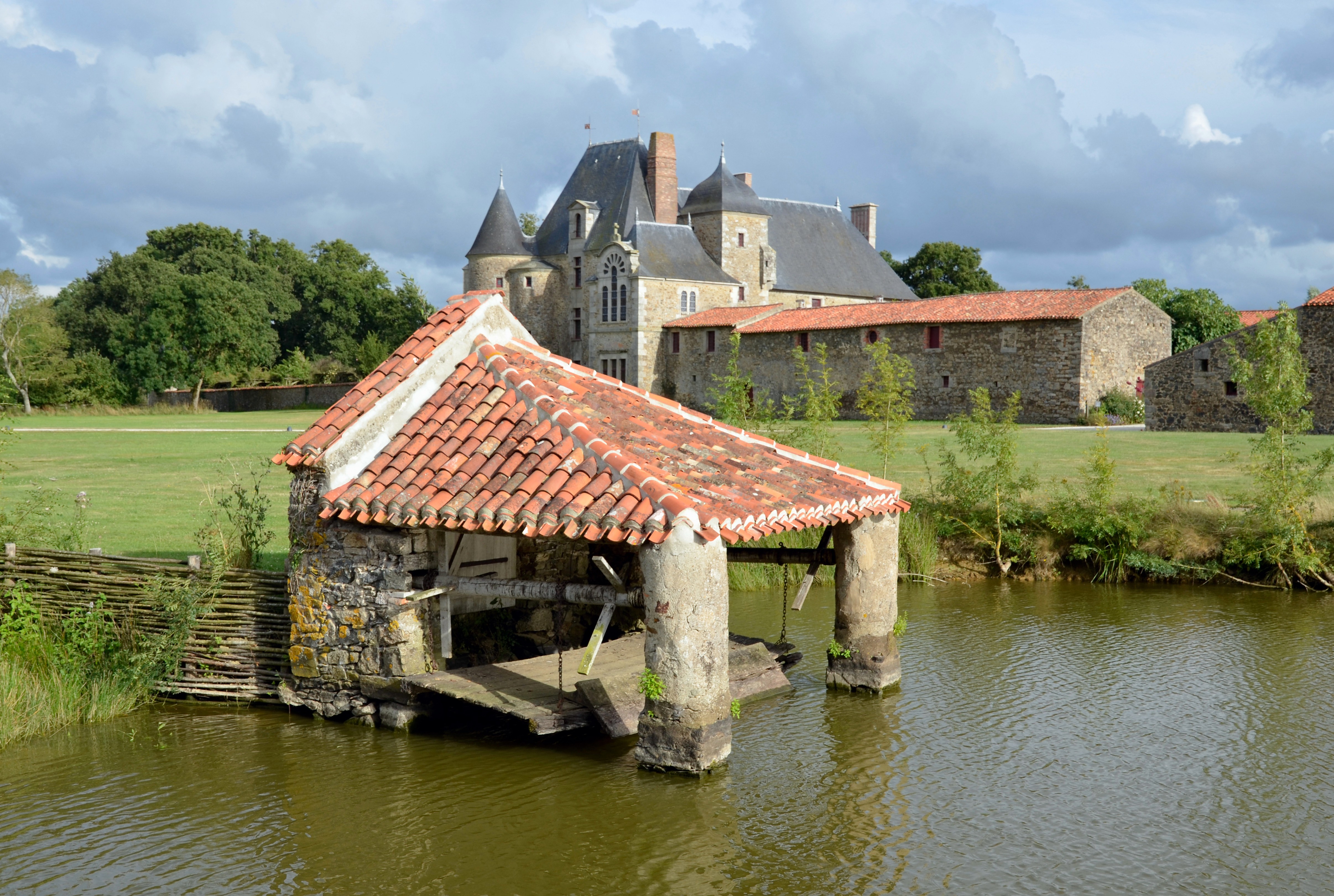 Vue depuis un lavoir au bord de l’eau sur le château de la Chabotterie et une partie du logis en pierre, entourés d’une pelouse d’herbe bien verte.
