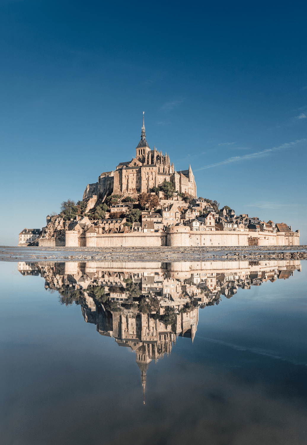 Reflet dans l'eau calme du Mont-Saint-Michel, abbaye médiévale sur un promontoir orcheux. Ciel bleu dégagé. Architecture historique. Murs en pierre. Toits pittoresques. Remparts. Symétrie. Sérénité