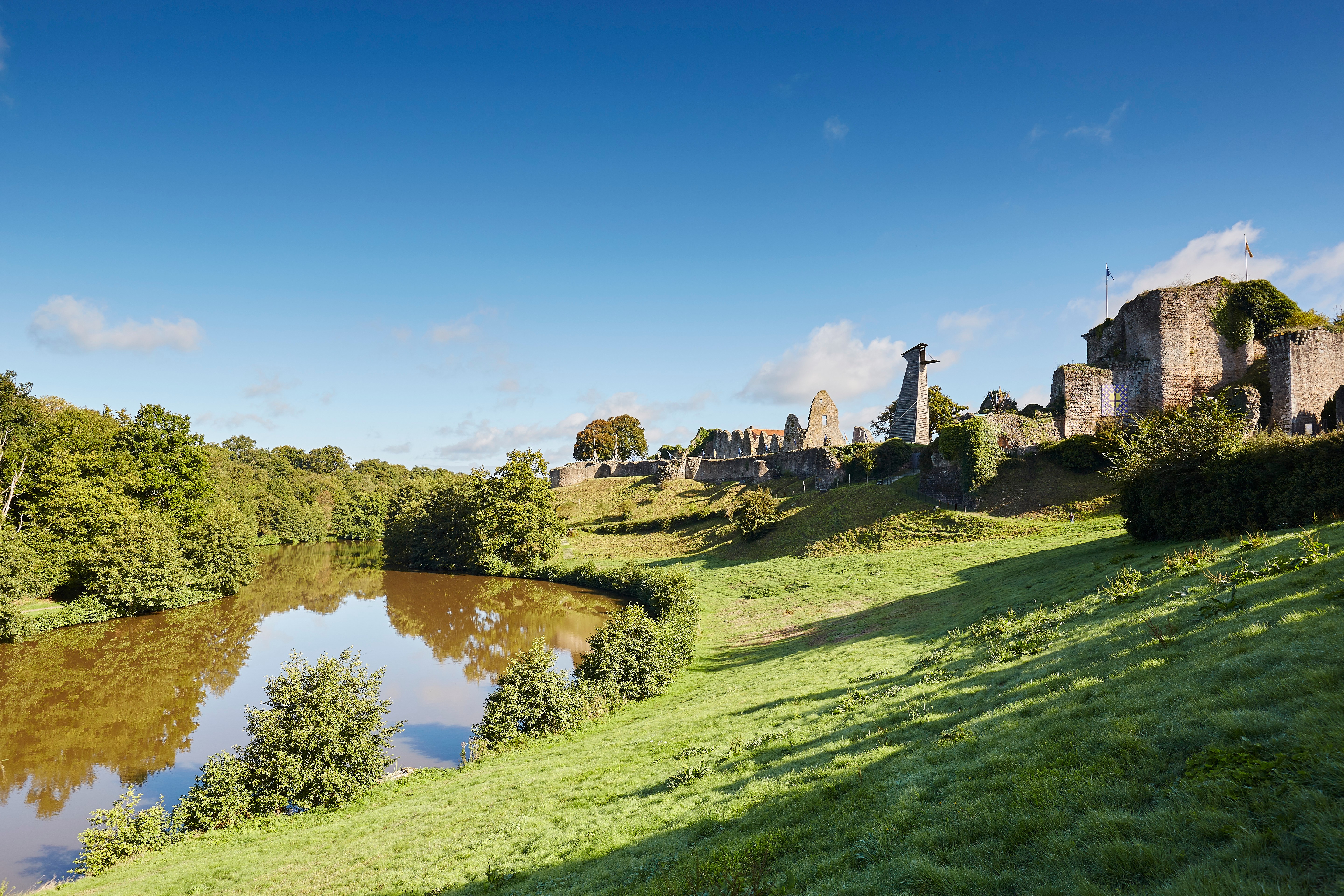 Vue sur un pré d'herbe verdoyant entre le ruisseau de la Crûme bordé d’arbres et les ruines du Château de Tiffauges 